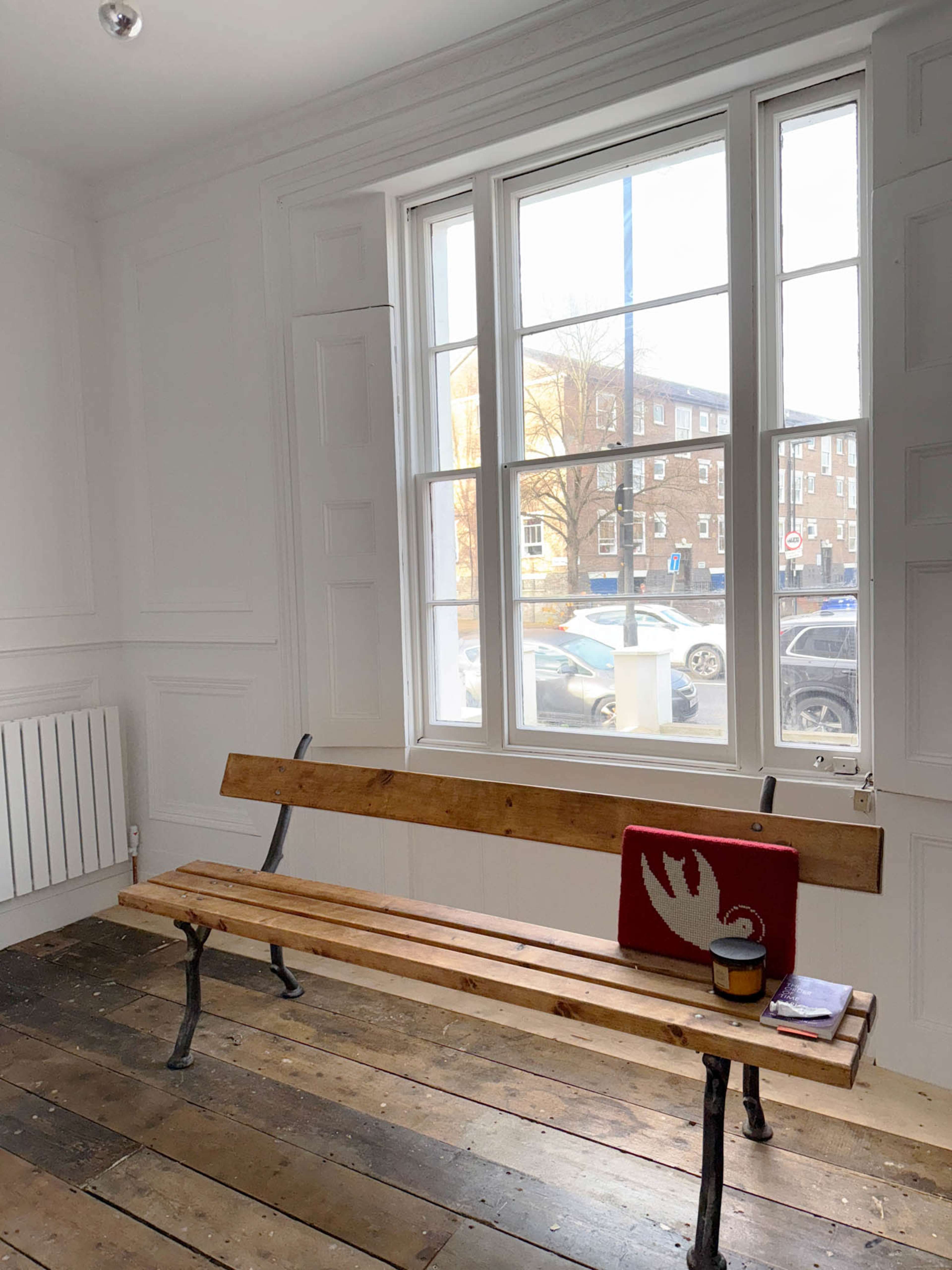 The image shows a wooden bench positioned near a large window in a room with plain white walls and a wooden floor, with a mug and a red item on the bench.