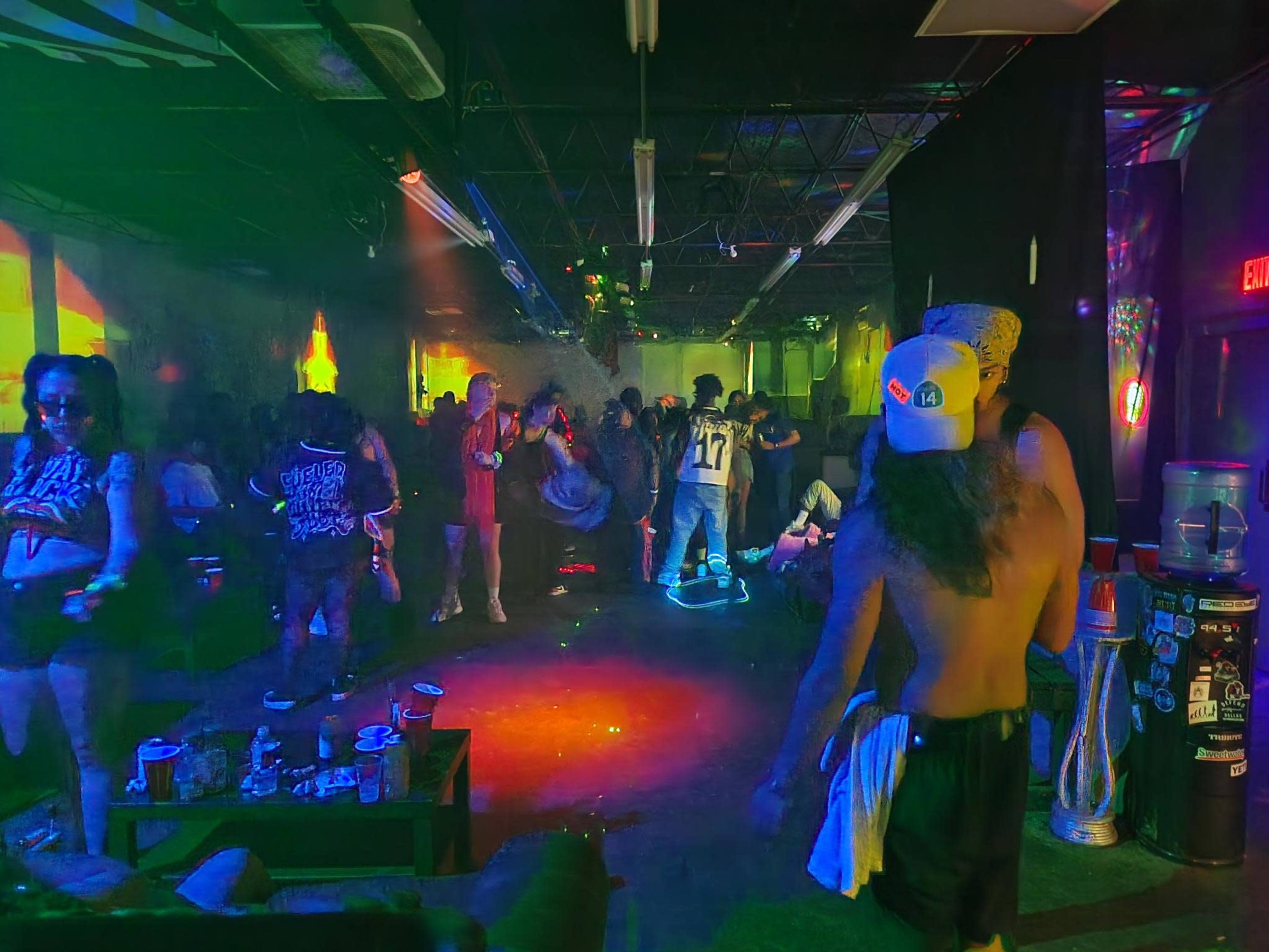 A lively indoor space features people dancing under colorful lights, with scattered furniture and a casual party atmosphere.