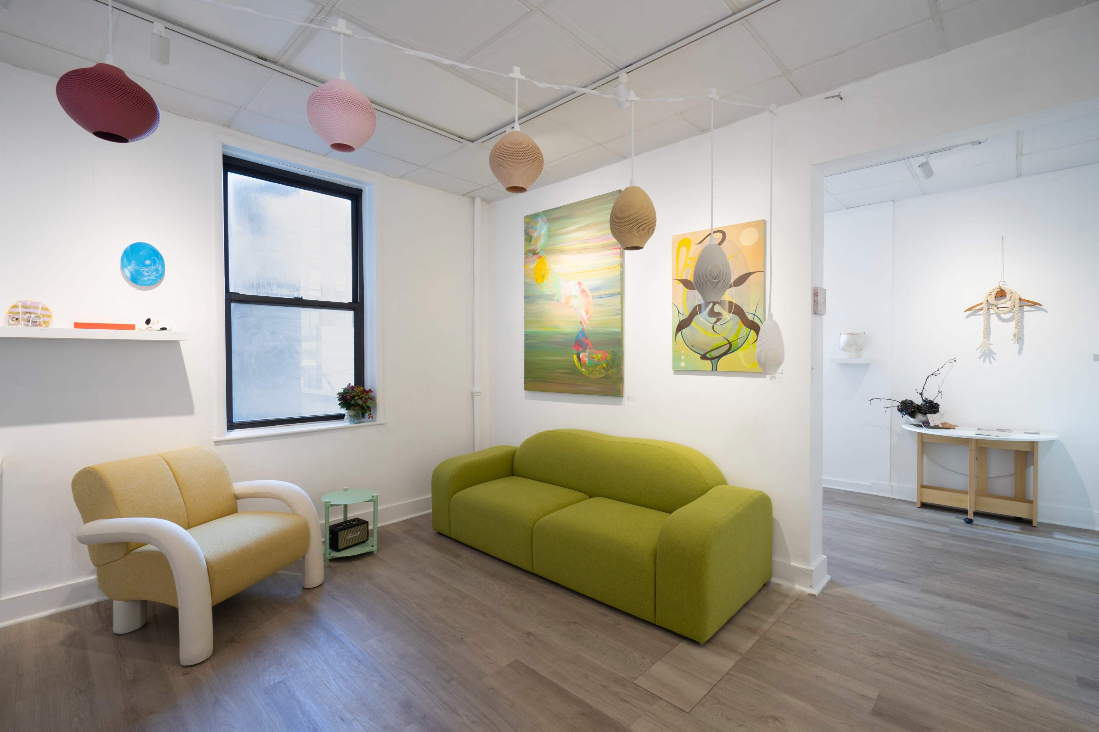 The image shows a modern art gallery featuring a green sofa, a light-colored armchair, various artworks on the walls, and soft pendant lighting.