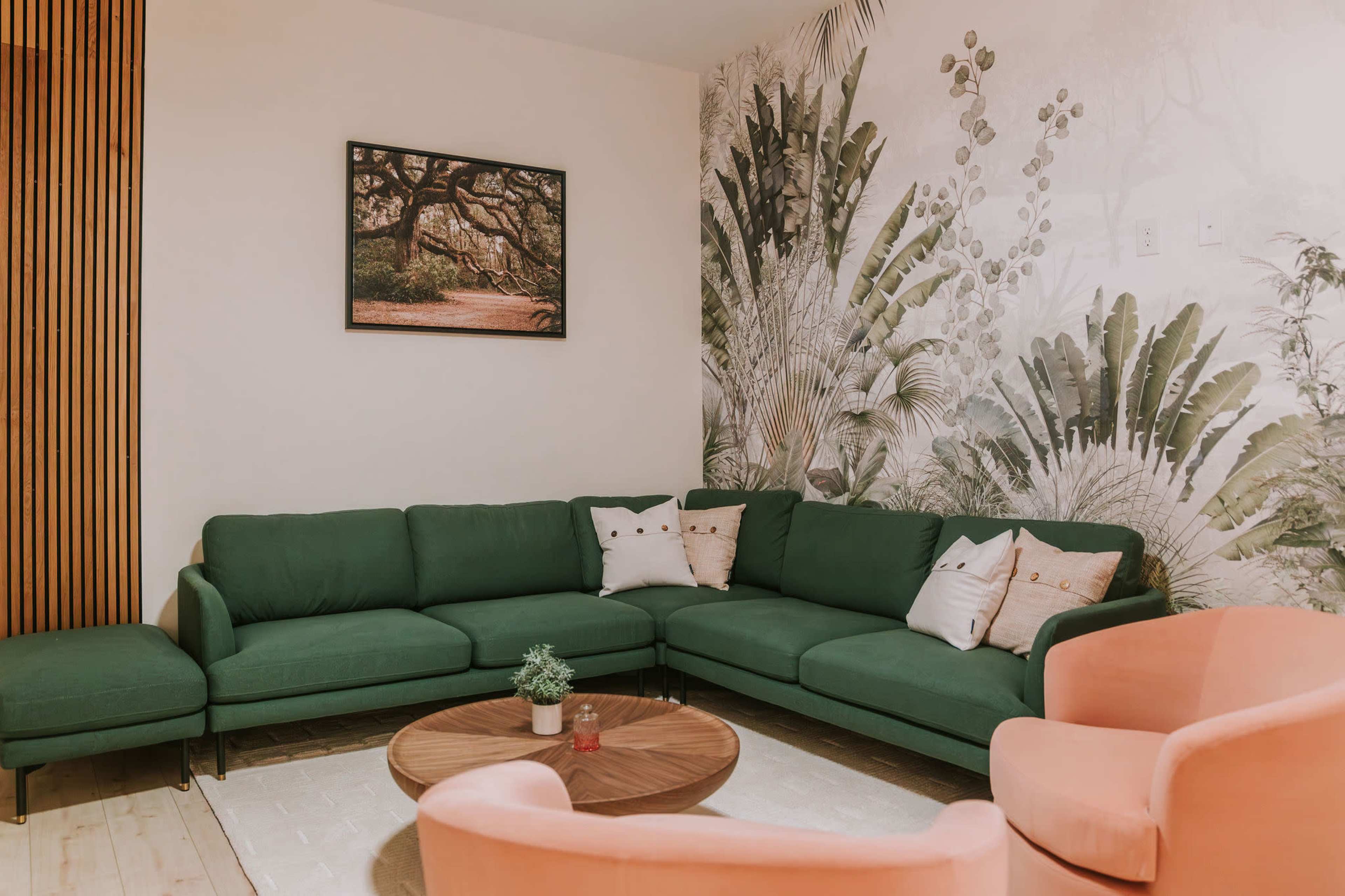 The image shows a modern living room featuring a large green sectional sofa, two light pink armchairs, a round wooden coffee table, and a wall adorned with a botanical wallpaper and a framed landscape photograph.