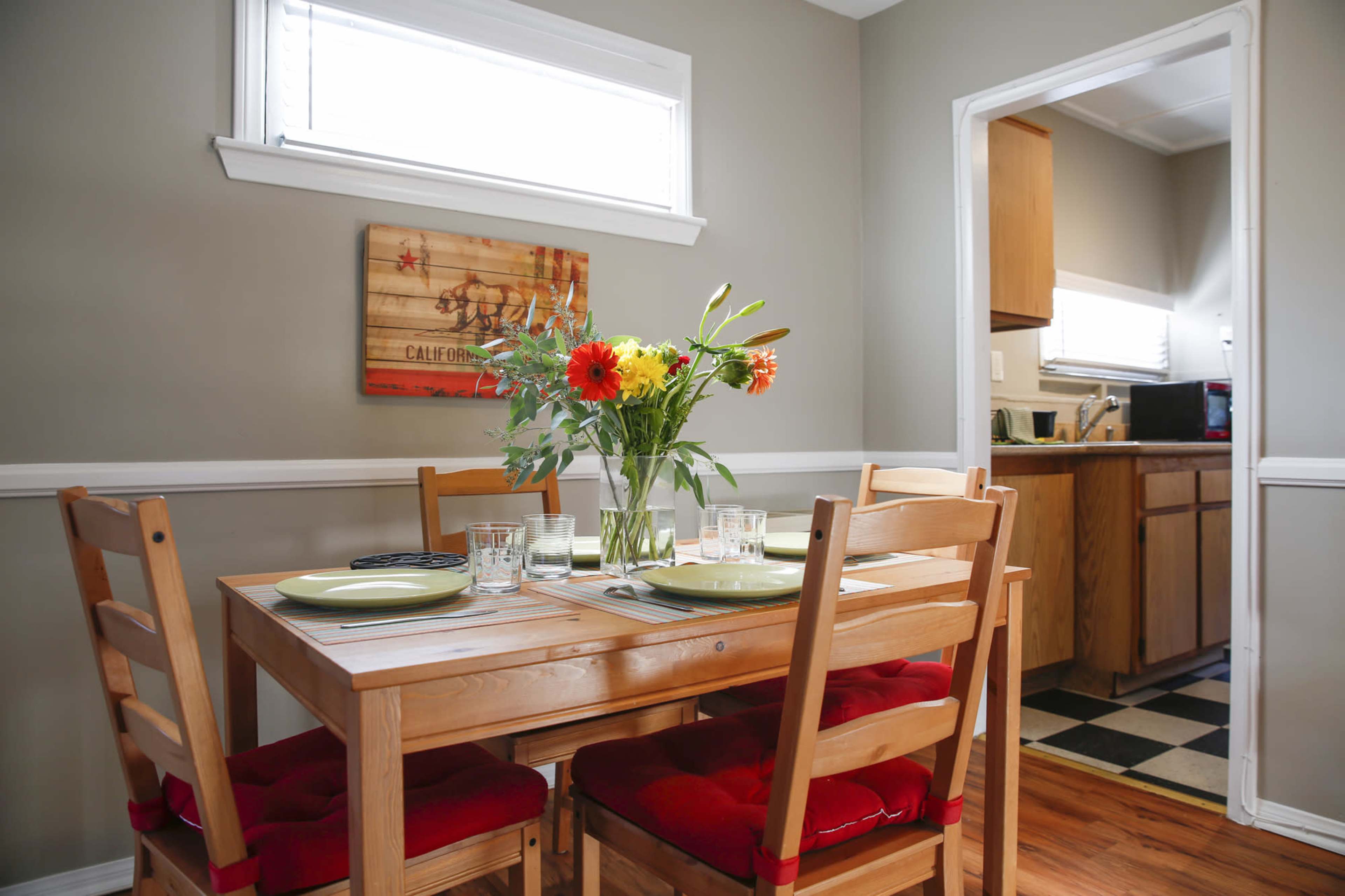 A wooden dining table with green plates and a vase of flowers is set in a kitchen-dining area featuring gray walls and a doorway leading to the kitchen.