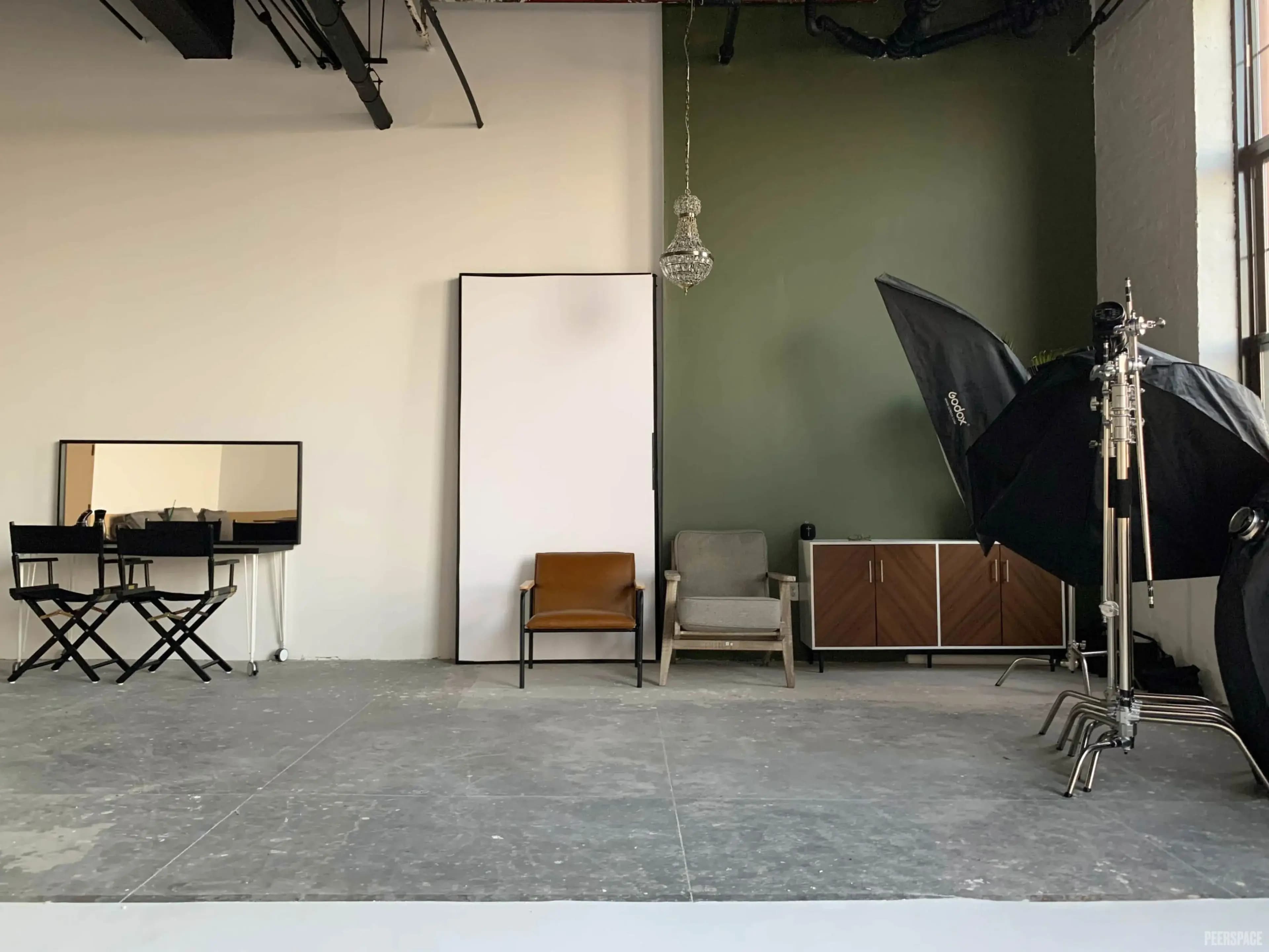 The image shows a photography studio with a green accent wall, a large backdrop, two chairs, a mirror, and lighting equipment arranged around the space.