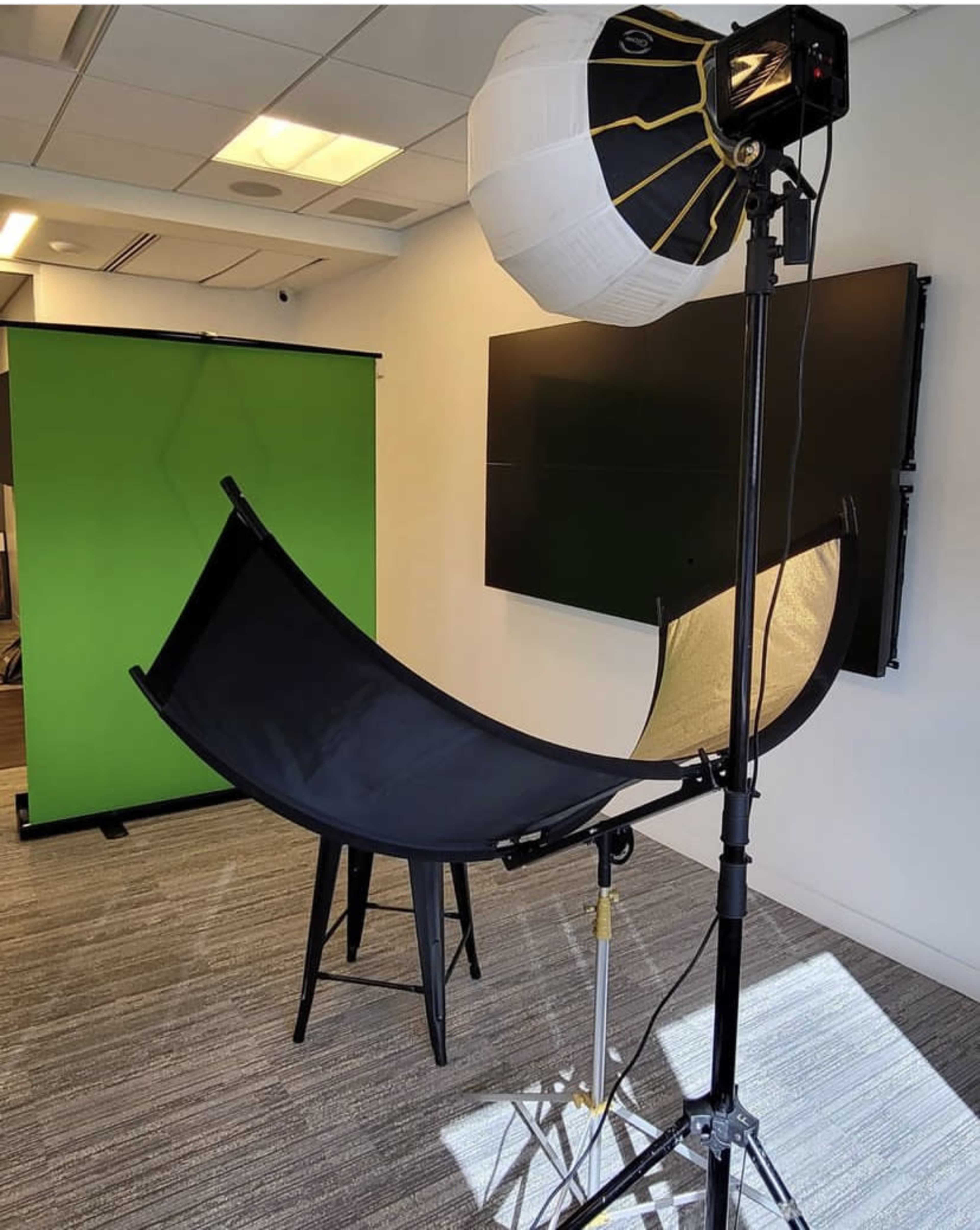 A photography setup featuring a black curved reflector, a light fixture with a softbox, and a green backdrop in an office environment.