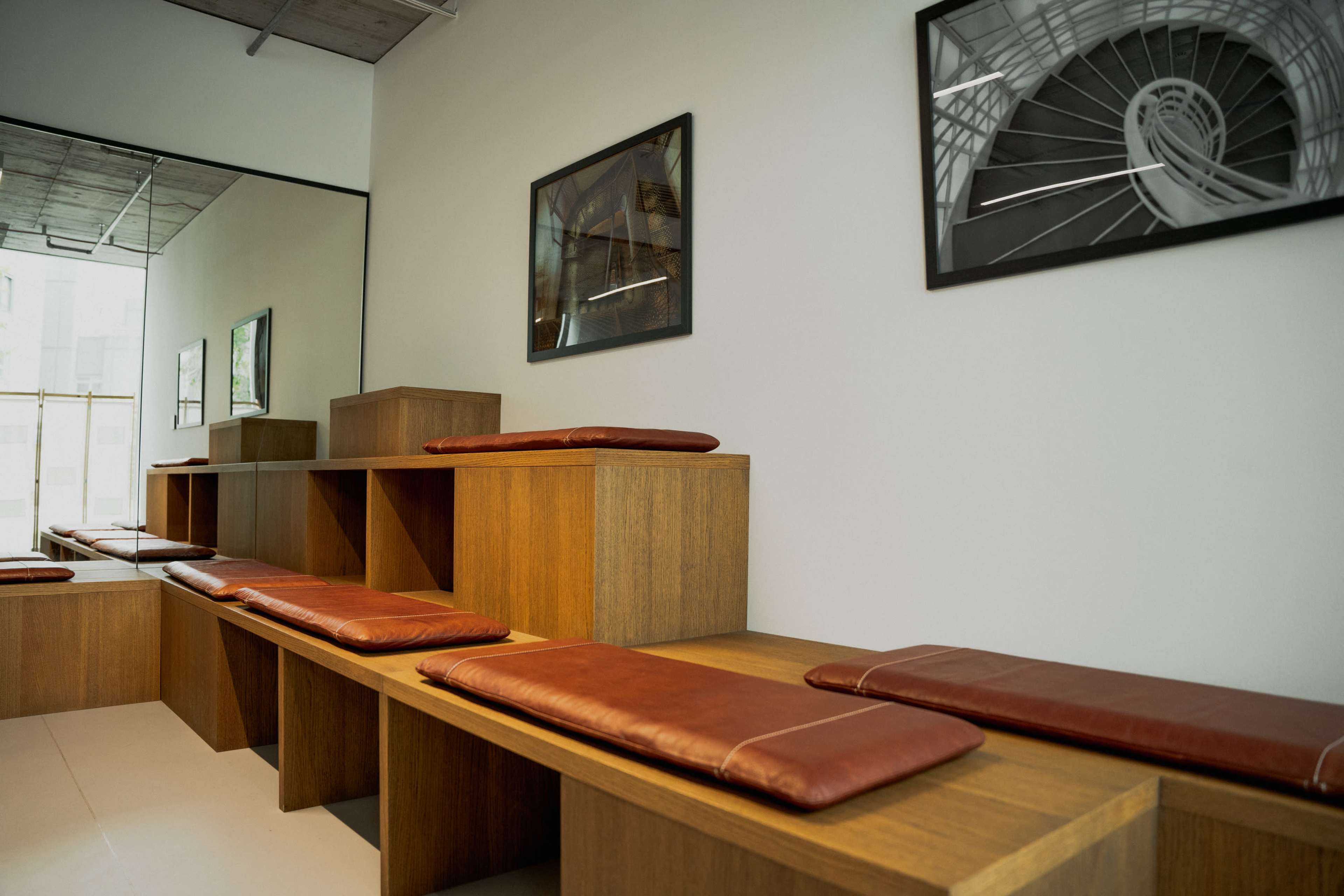 The image depicts a minimalist interior space with wooden seating and brown cushions, adorned with framed black-and-white photographs on the walls.