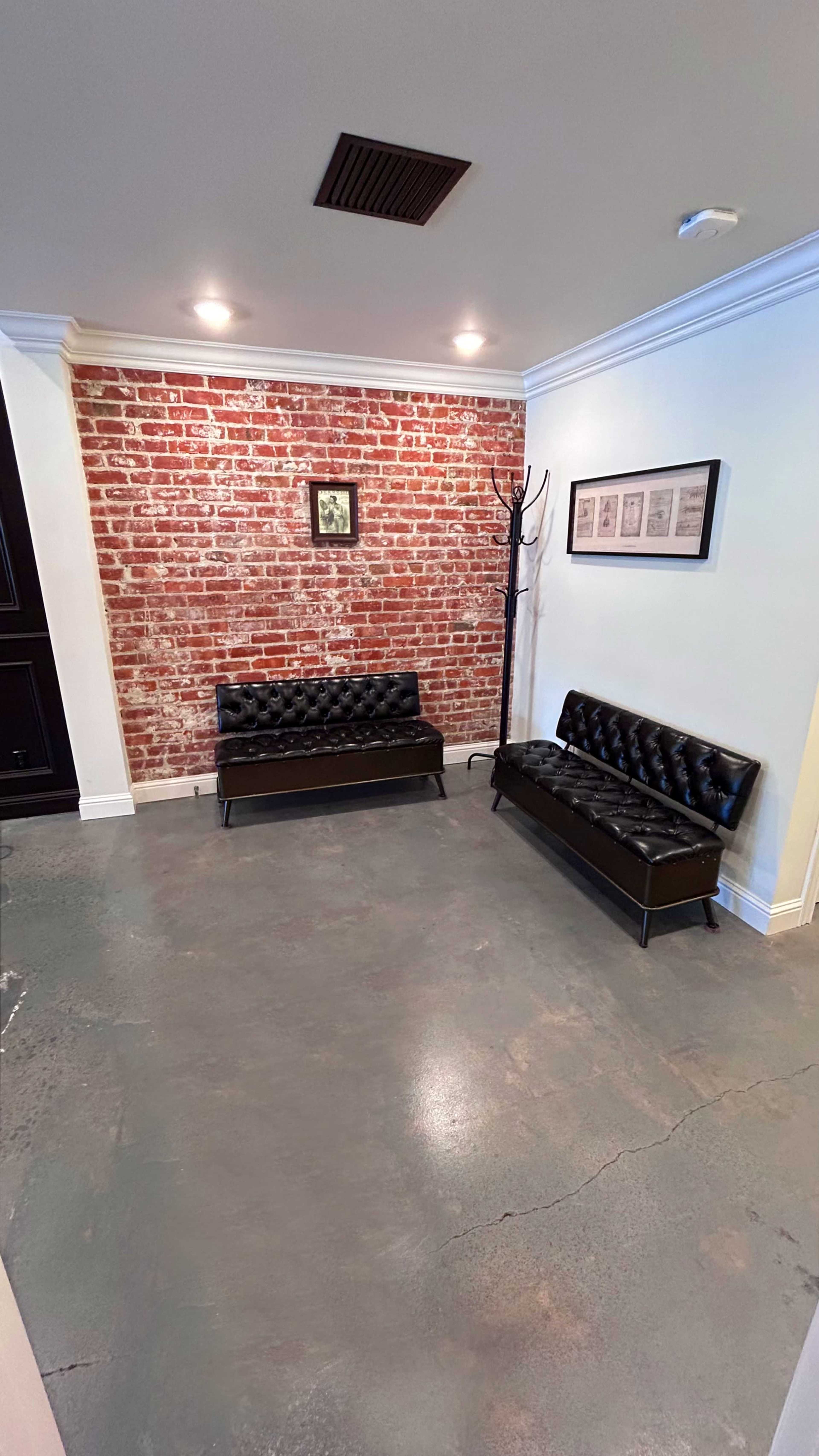 A minimalist room with two black leather couches facing a bare brick wall, accompanied by a coat rack and framed pictures.