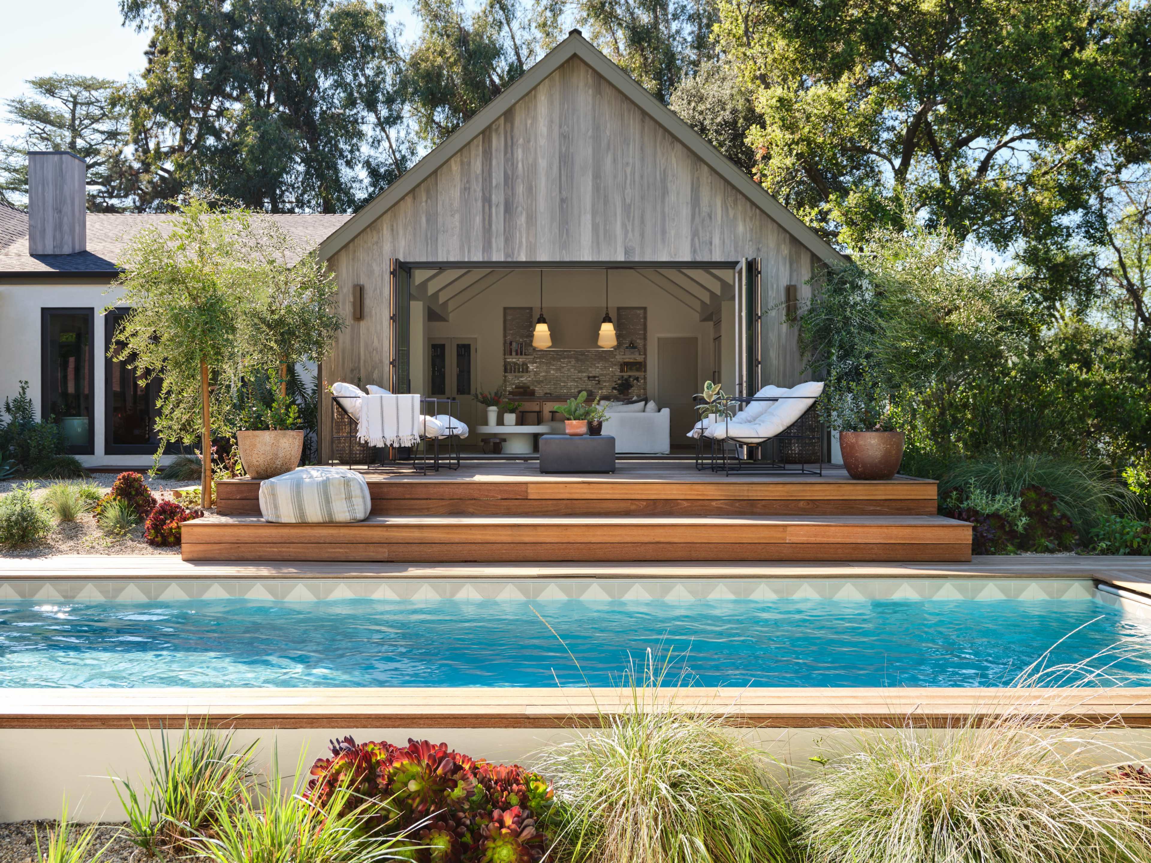 A large wooden deck with seating overlooks a swimming pool, adjacent to a modern house framed by trees.