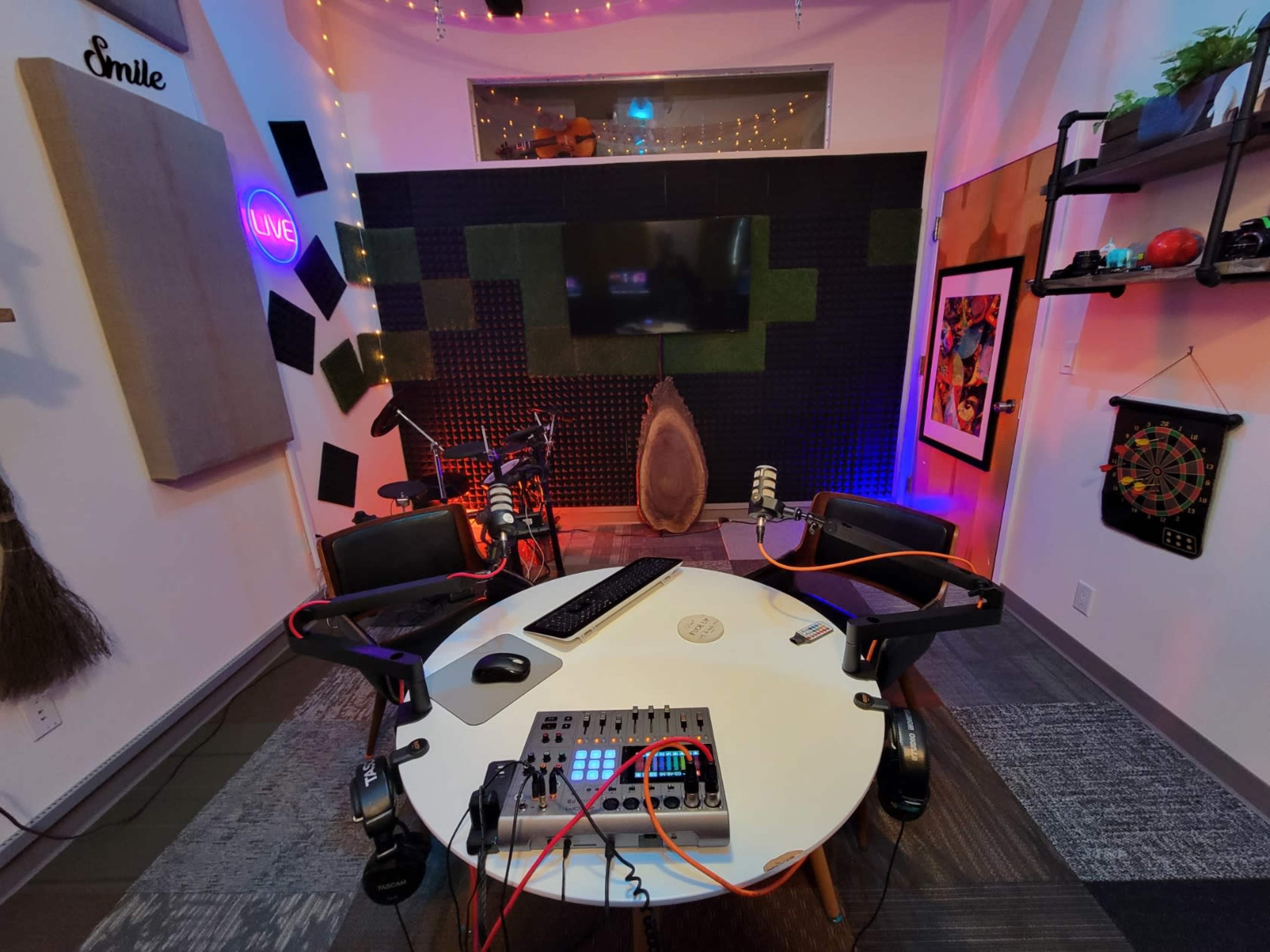 A recording studio setup featuring a circular table with microphones, a computer, and colorful lighting against a backdrop of soundproofing panels and a large screen.