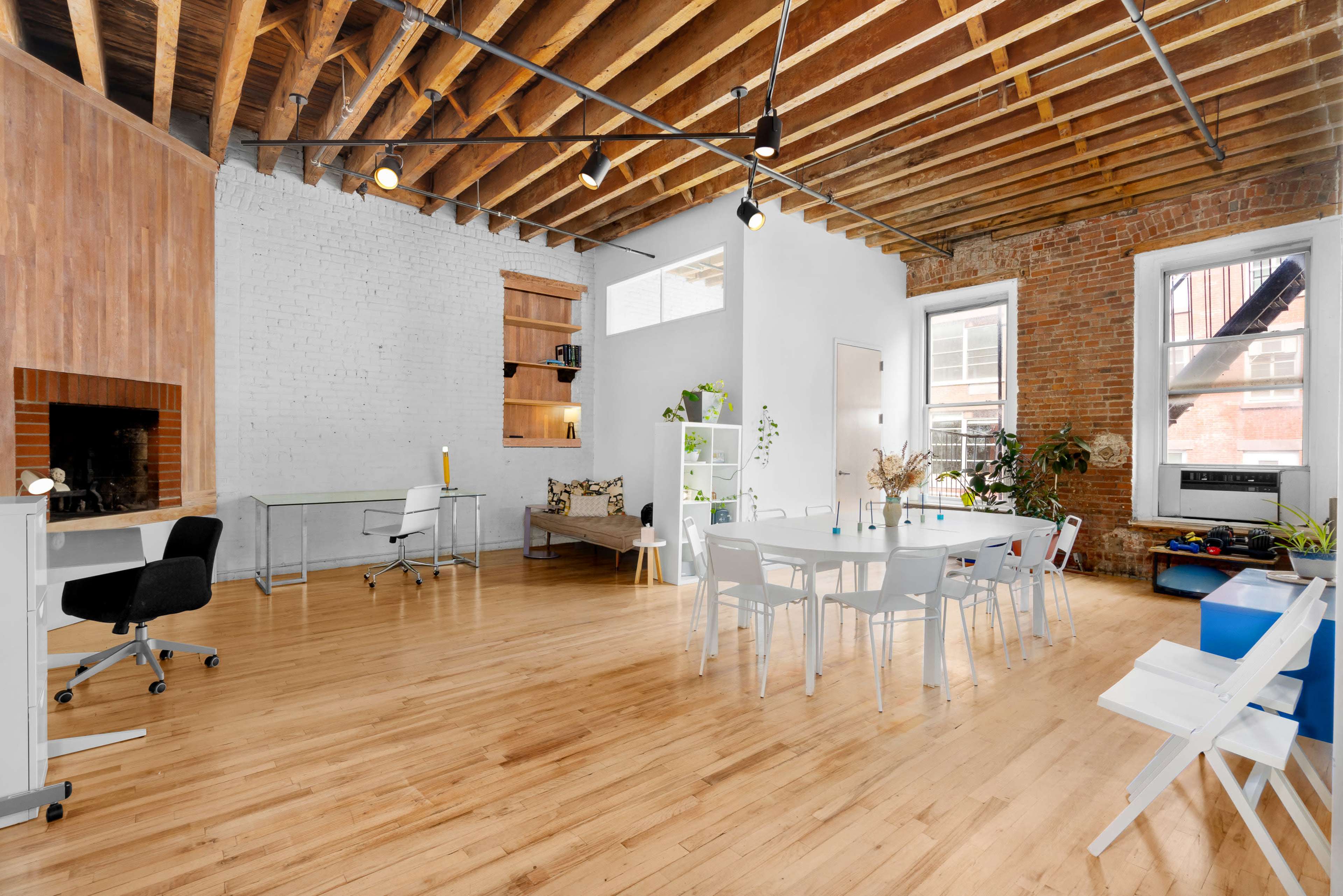 A spacious, modern interior featuring wood flooring, exposed beams, and a bright seating area with a dining table, workstations, and decorative plants.