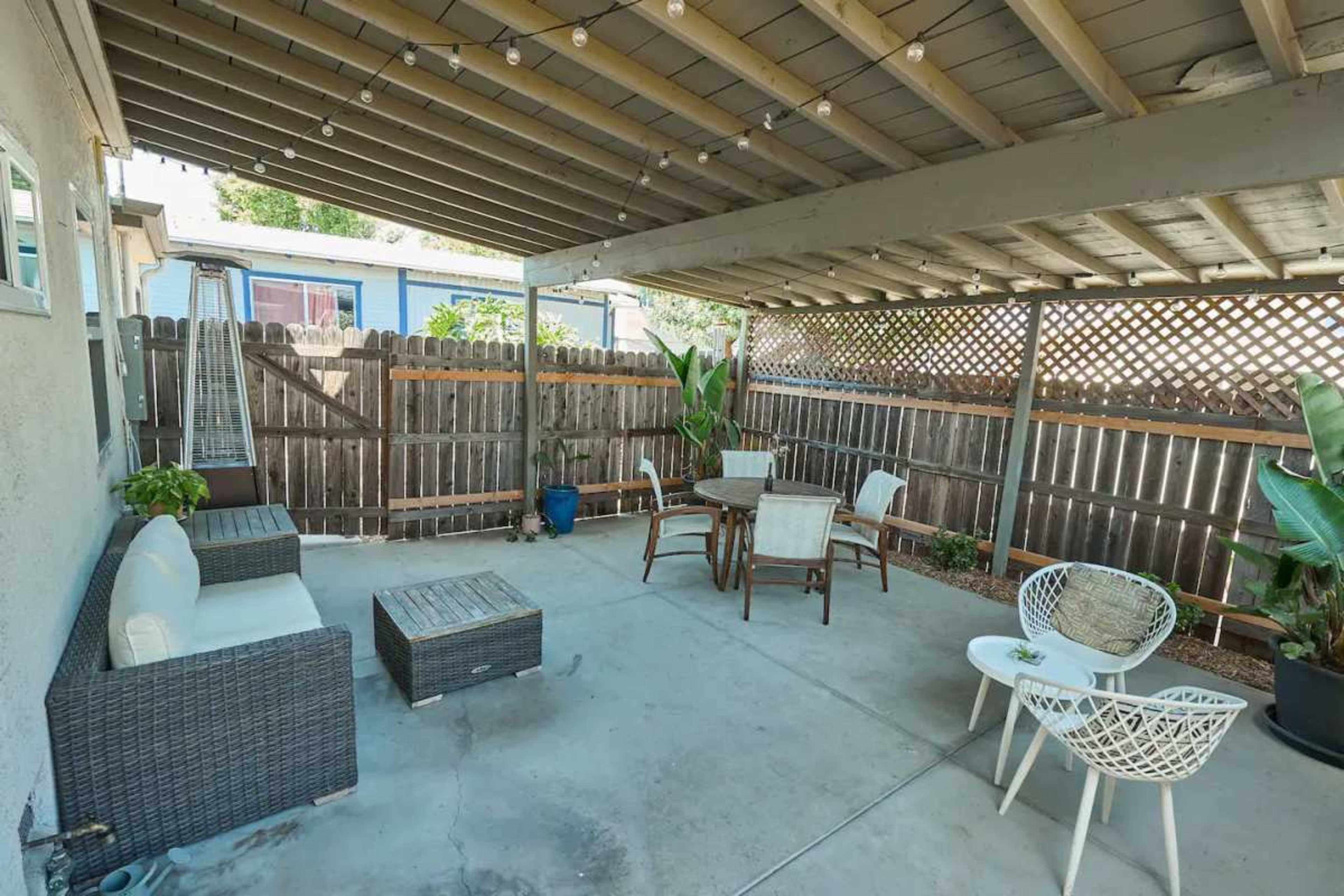 The image shows a spacious covered patio area with a seating arrangement, including a circular table and chairs, surrounded by a wooden fence and plants.