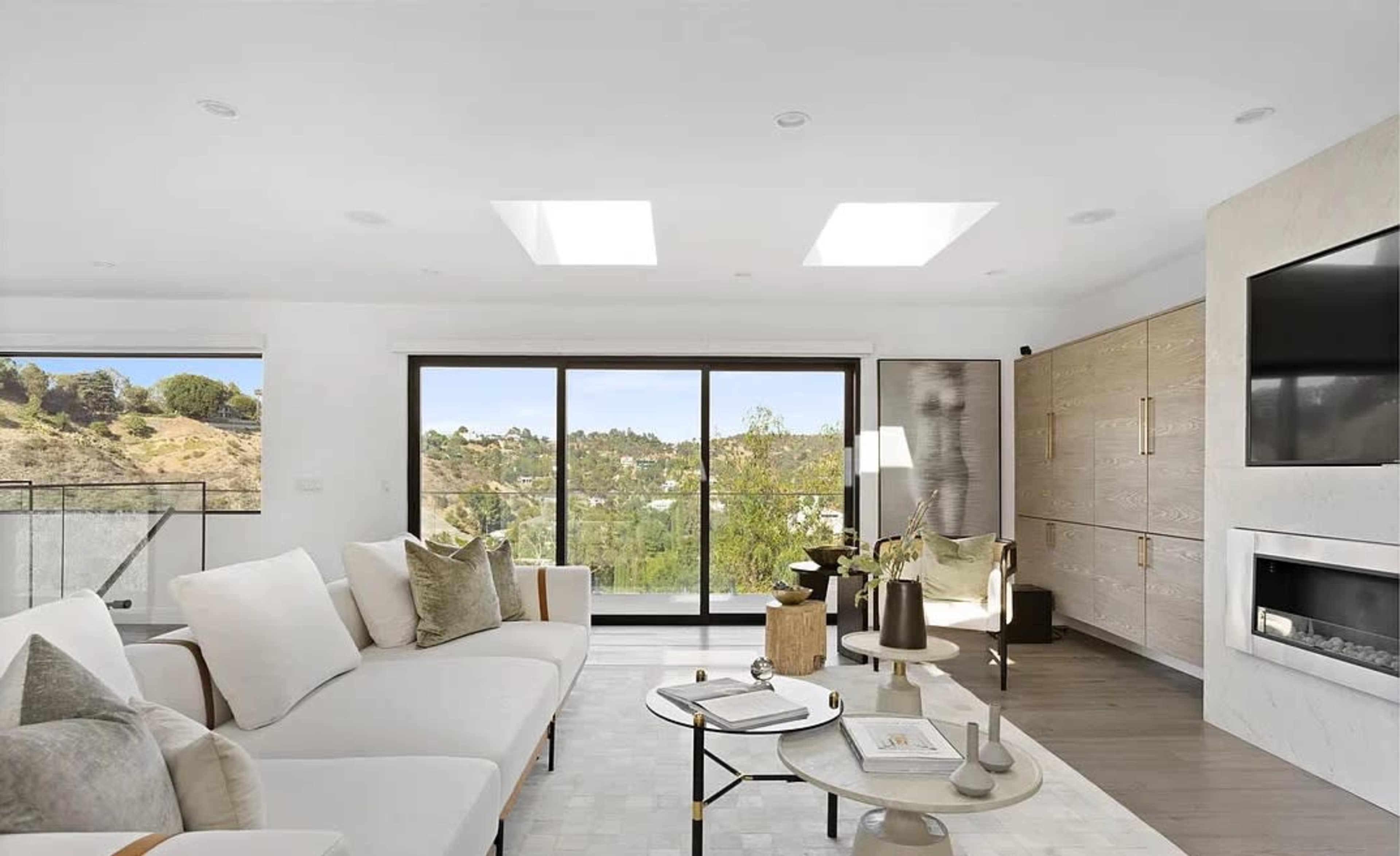 The image depicts a modern living room with large windows, a sectional sofa, a coffee table, and a wall-mounted television, overlooking a scenic view.