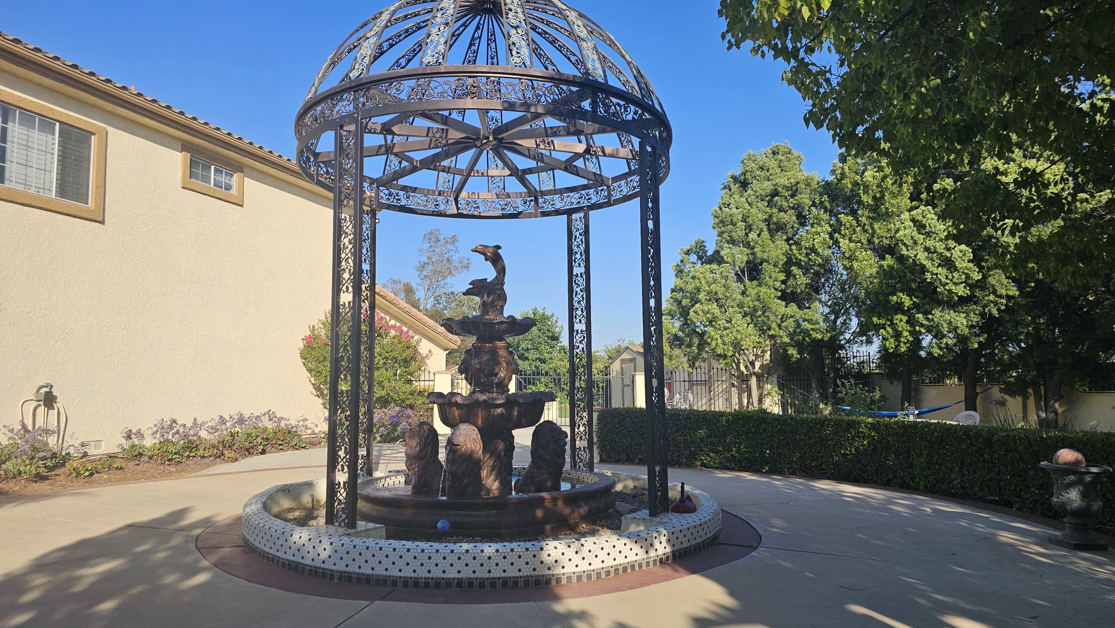 The image shows a decorative fountain encased in a domed metal structure, surrounded by landscaped greenery and a paved area.