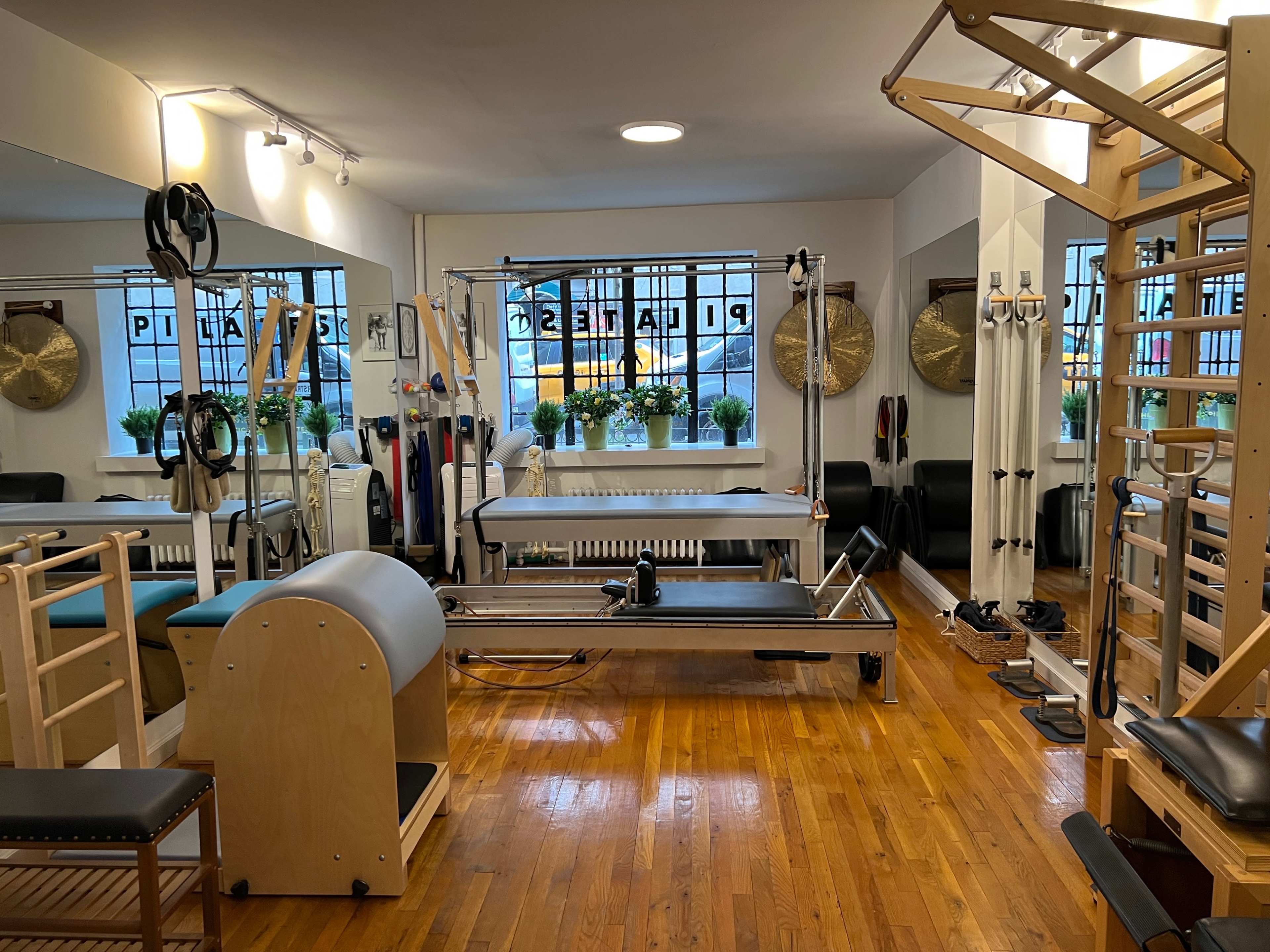 The image shows a well-equipped Pilates studio featuring various exercise machines, mirrors, and natural light coming through large windows.