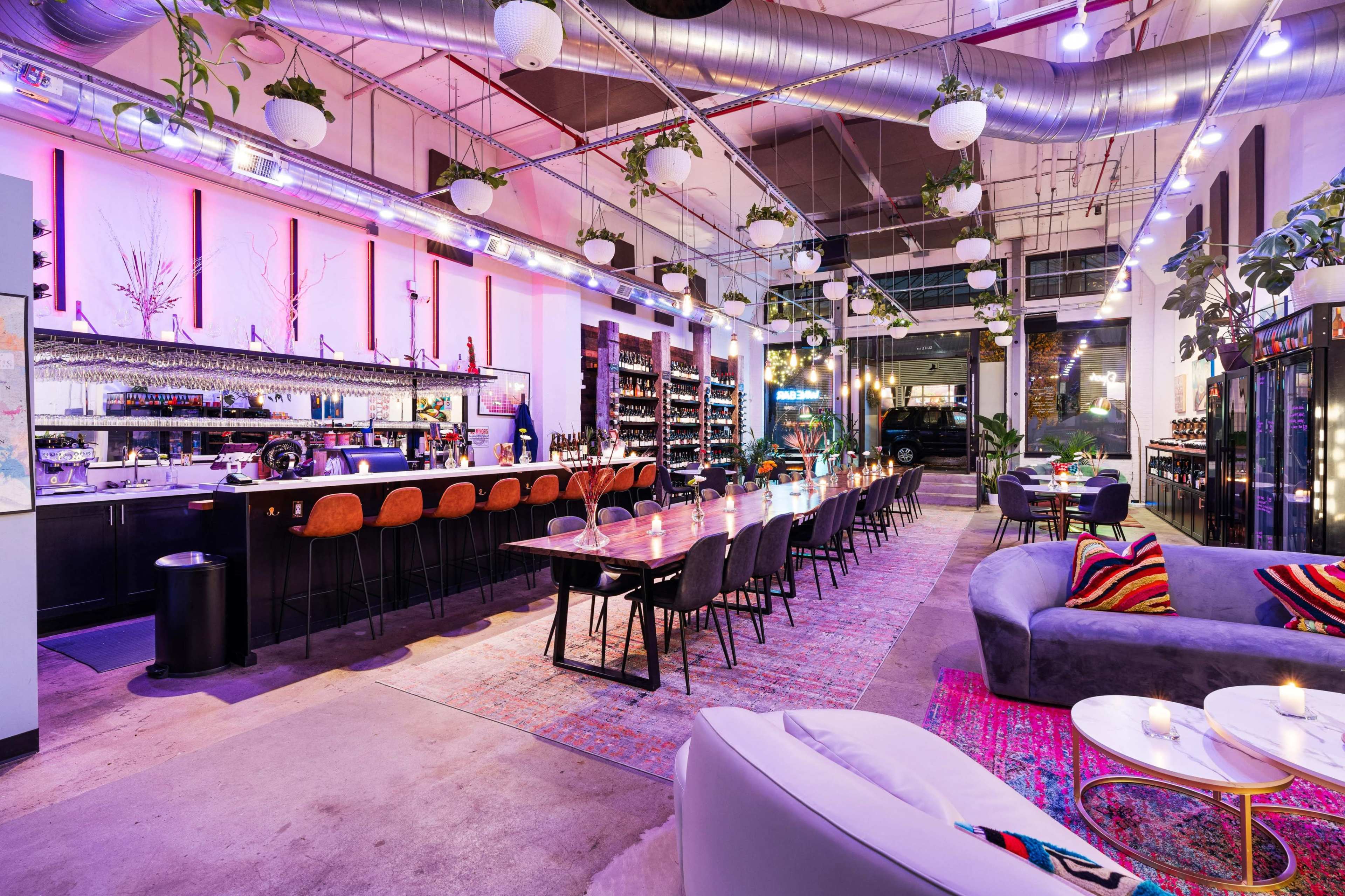 A modern restaurant interior with a long communal dining table, a bar area, and decorative plants hanging from the ceiling.