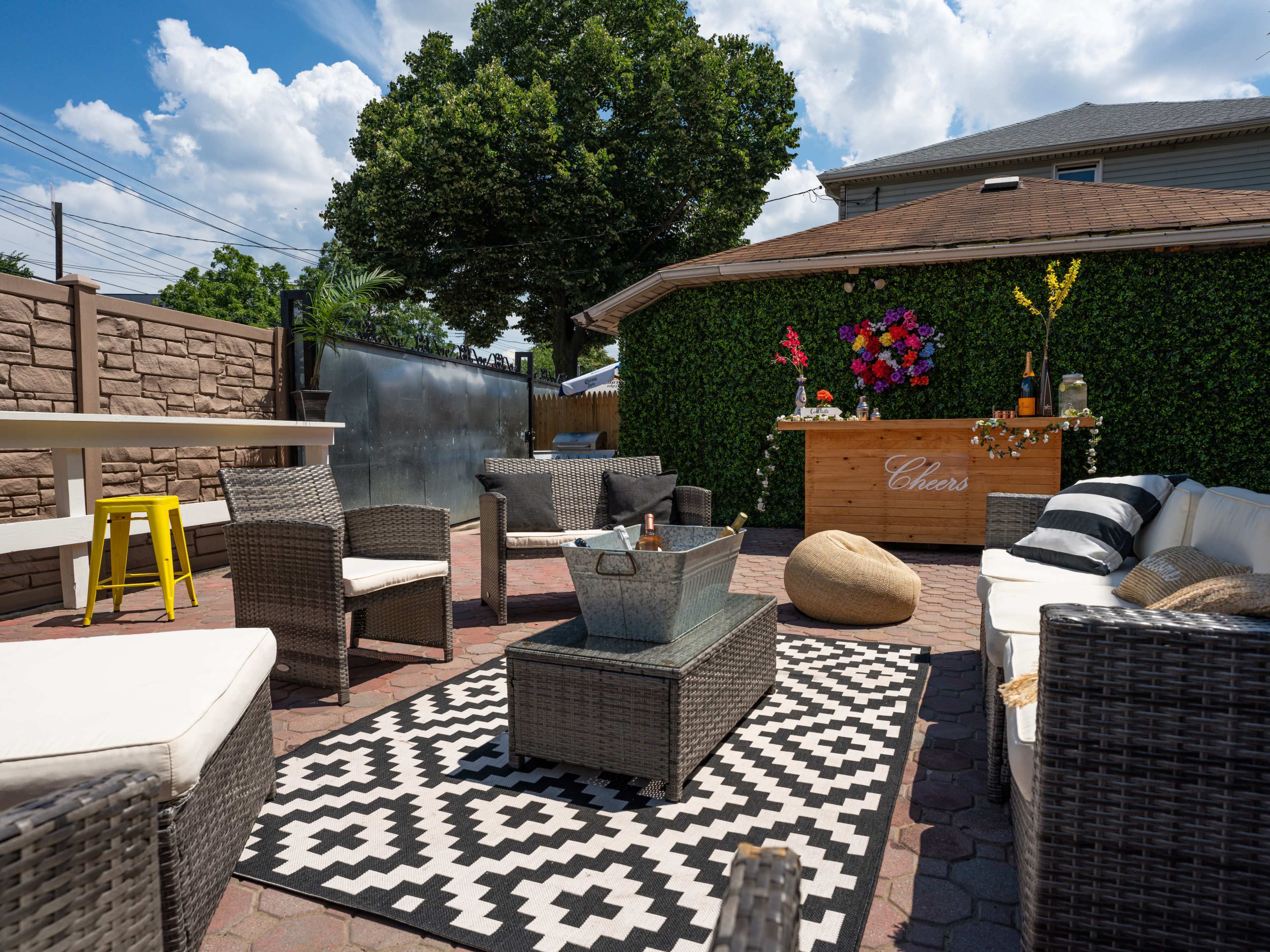 A patio area with seating arrangements, a patterned rug, a fire pit, and a bar decorated with flowers against a backdrop of green foliage.