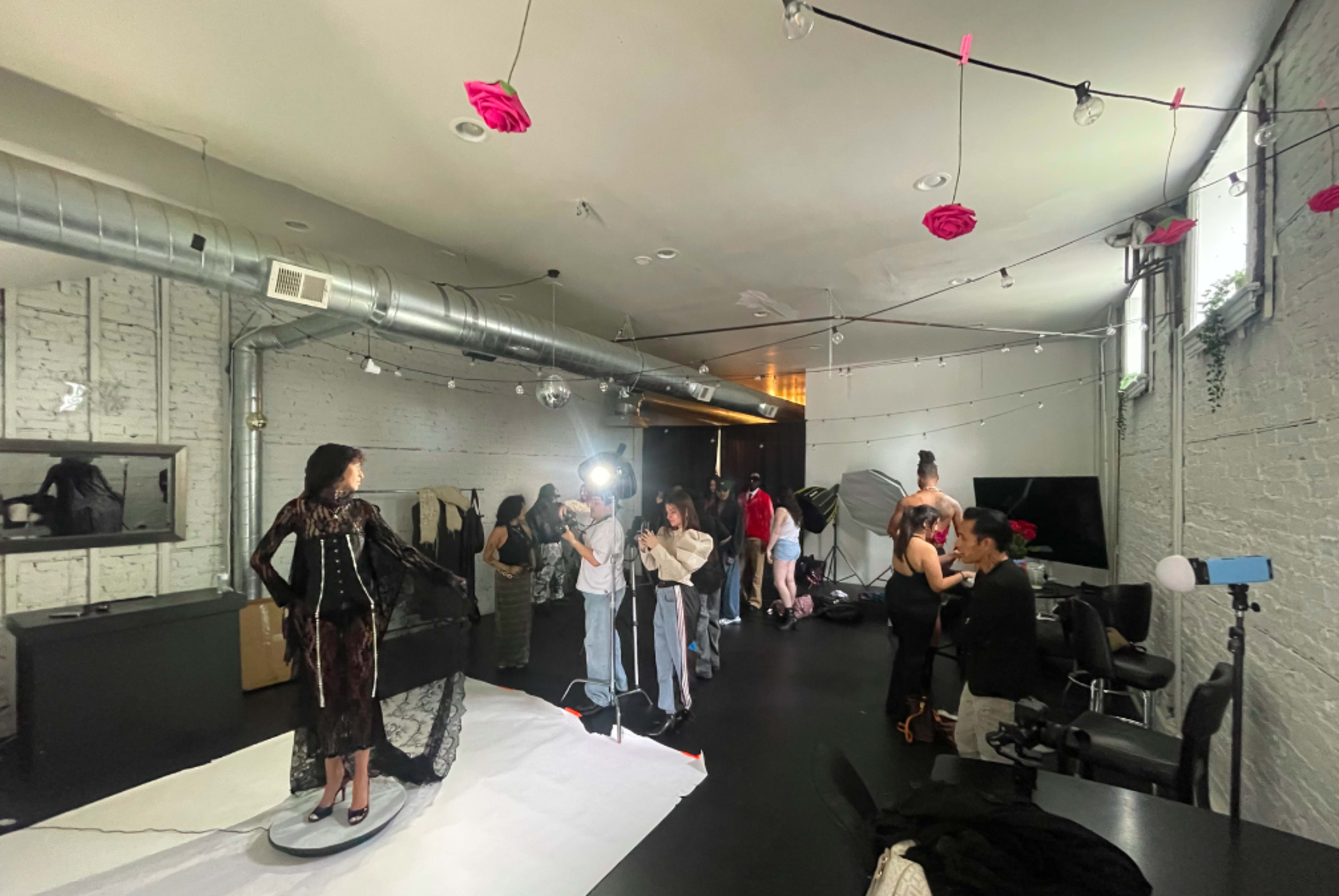 A fashion photoshoot is taking place in a studio, with models being styled and photographed under bright lights.