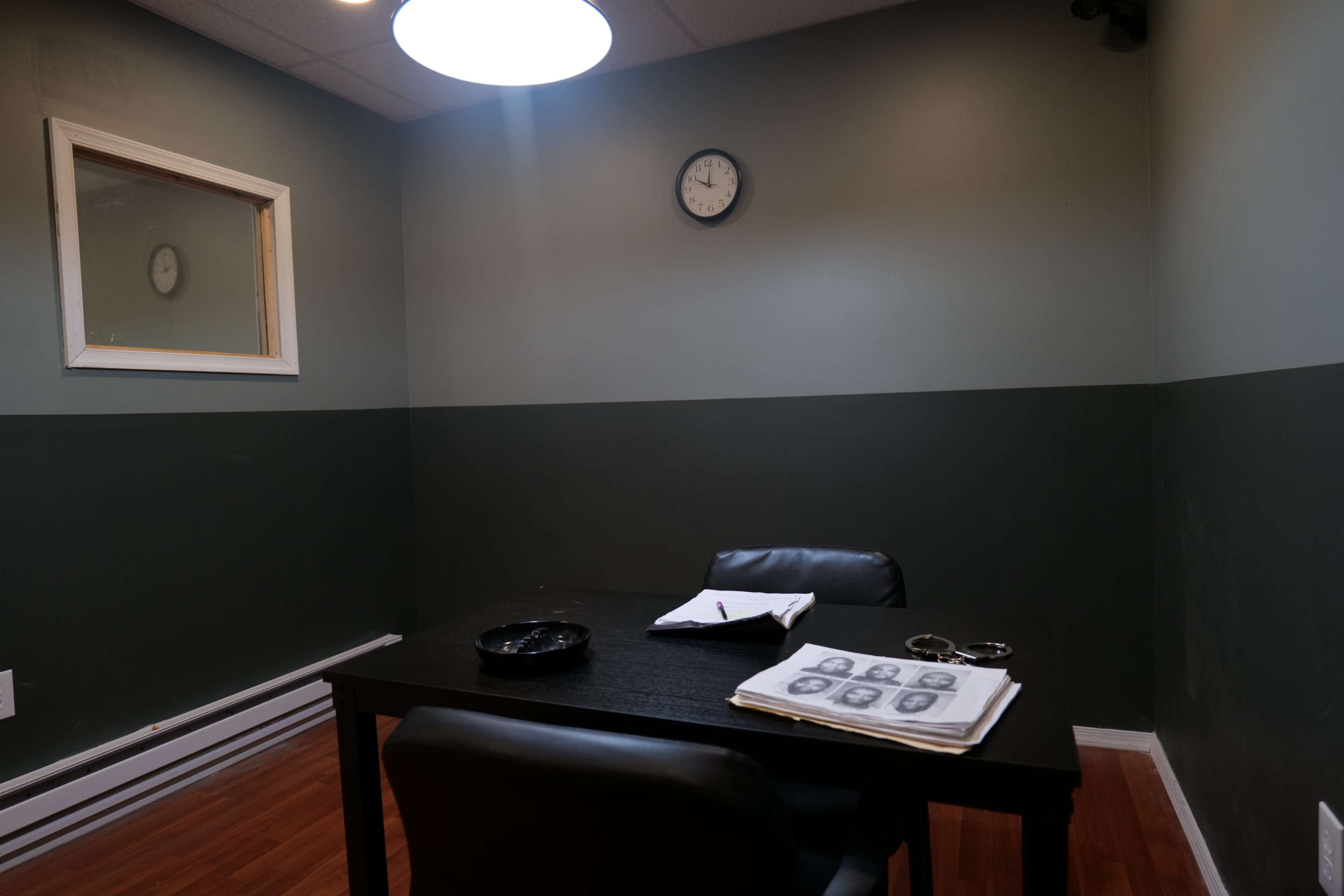 A small, dimly lit room with a table and two chairs, a clock on the wall, and a framed window, along with papers and an ashtray on the table.