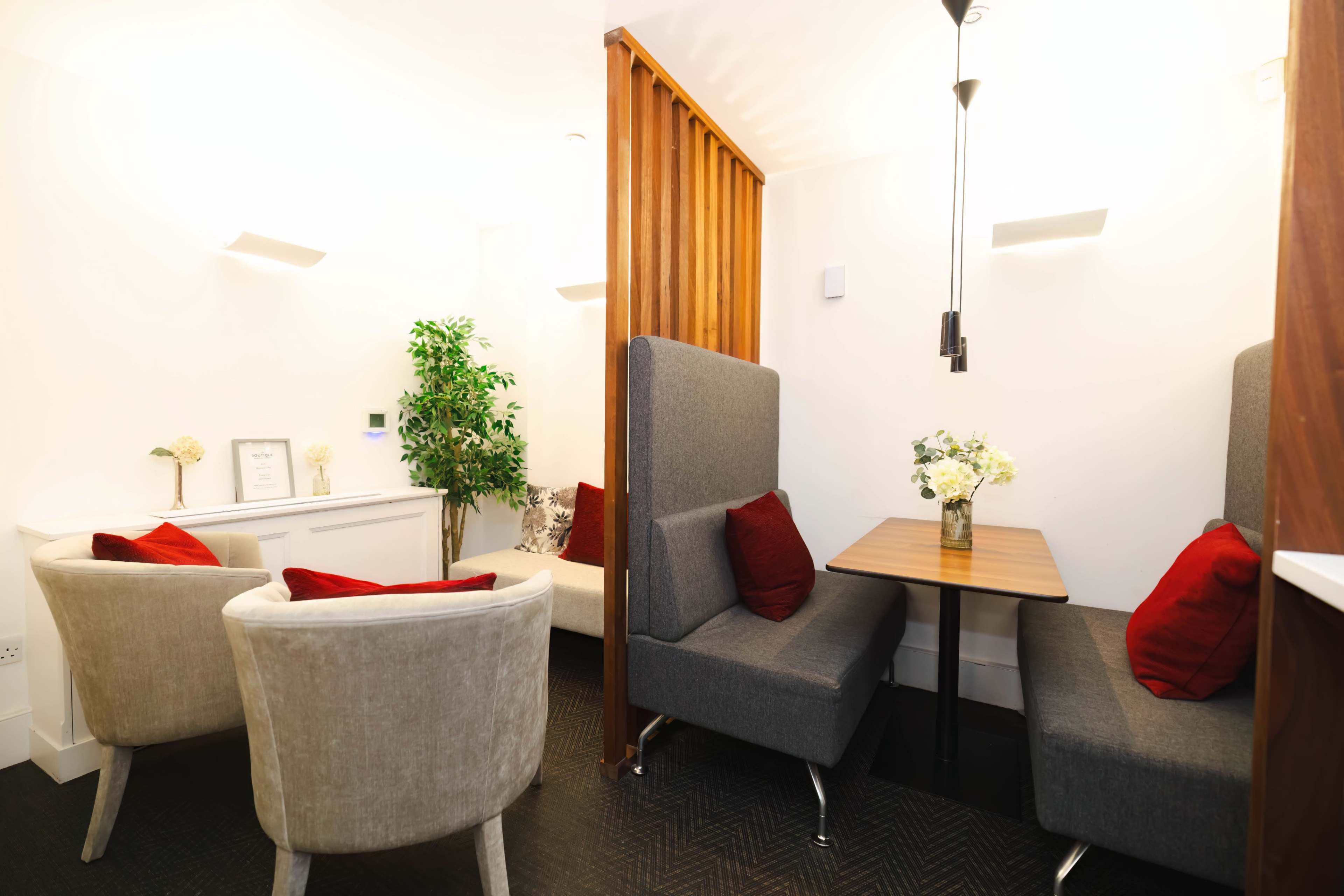 A small room features two grey armchairs with red cushions and a wooden table beside a partitioned seating area with a potted plant.