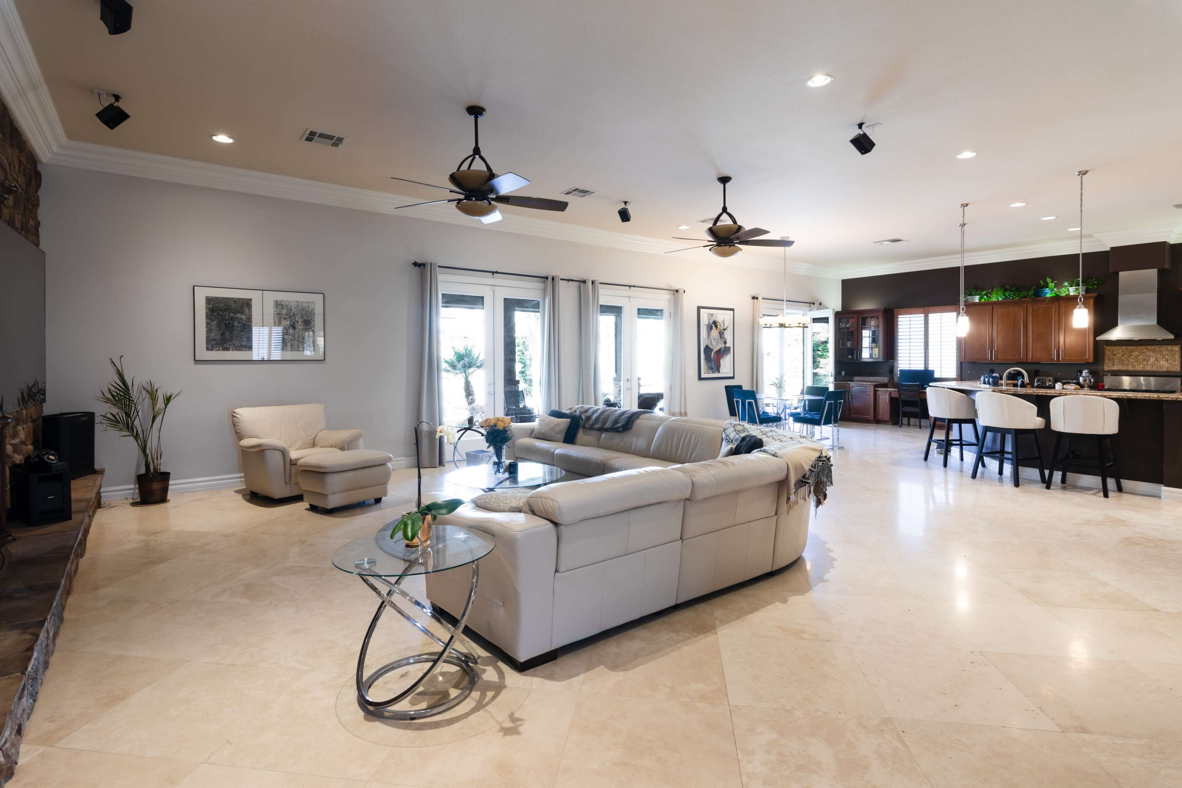 A spacious living room features a light-colored leather sectional sofa, a glass coffee table, and sliding doors that lead to an outdoor area, with a kitchen and dining space visible in the background.