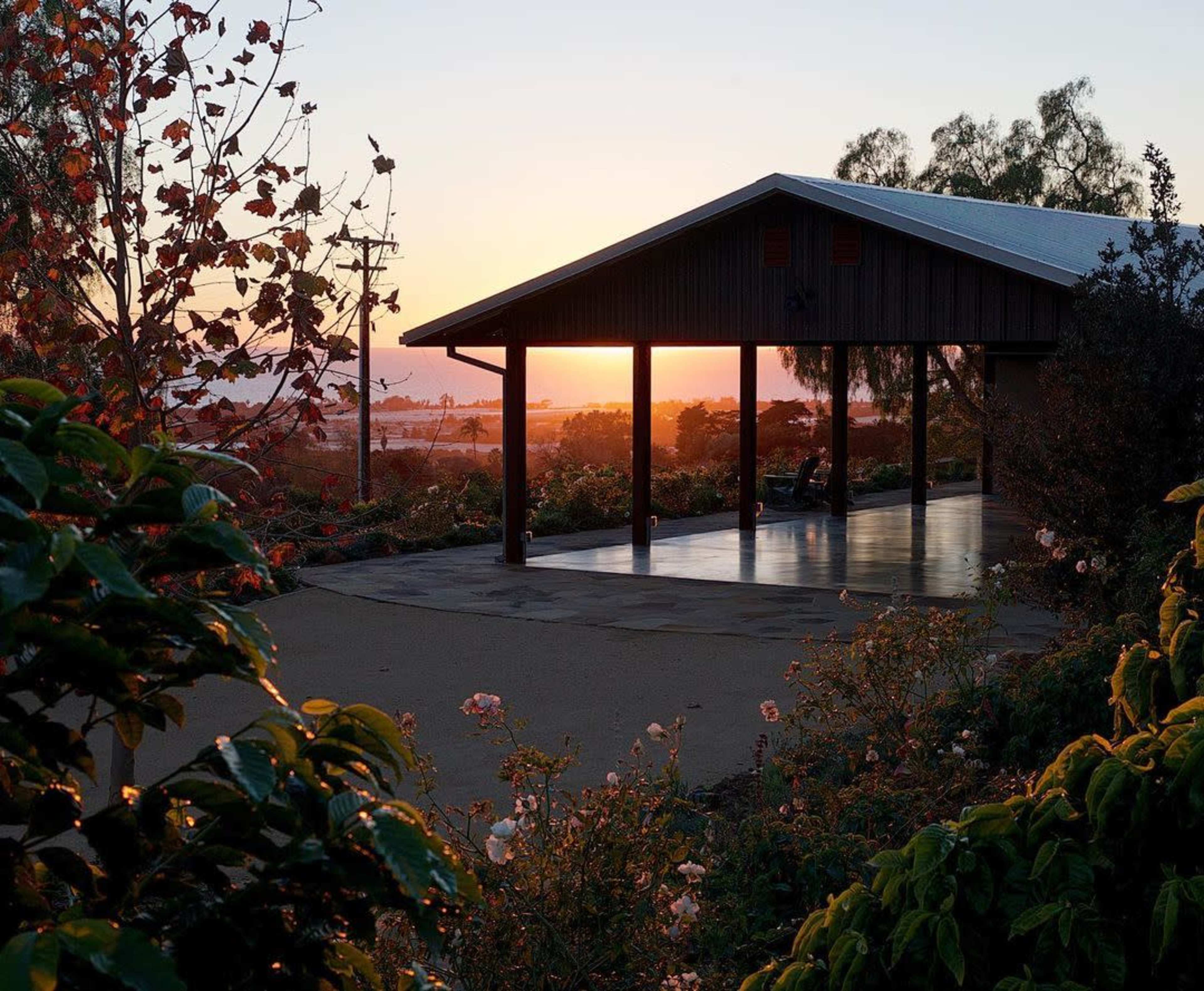 A modern wooden structure stands with open sides as the sun sets behind it, illuminating the surrounding garden and landscape.