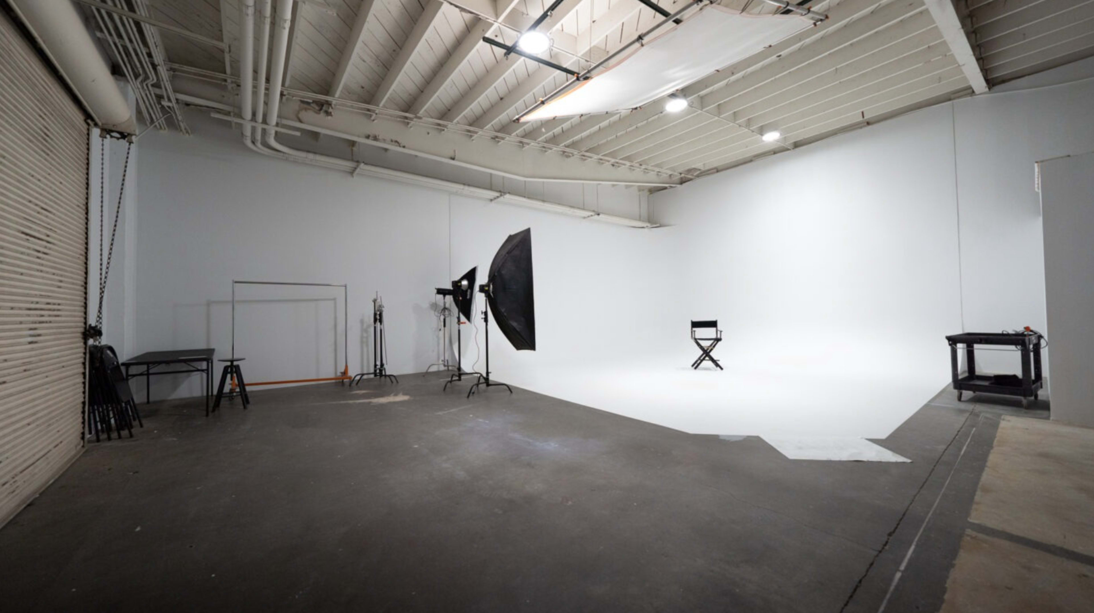 A spacious photography studio featuring a stark white backdrop, a folding chair, lighting equipment, and various supports along the walls.