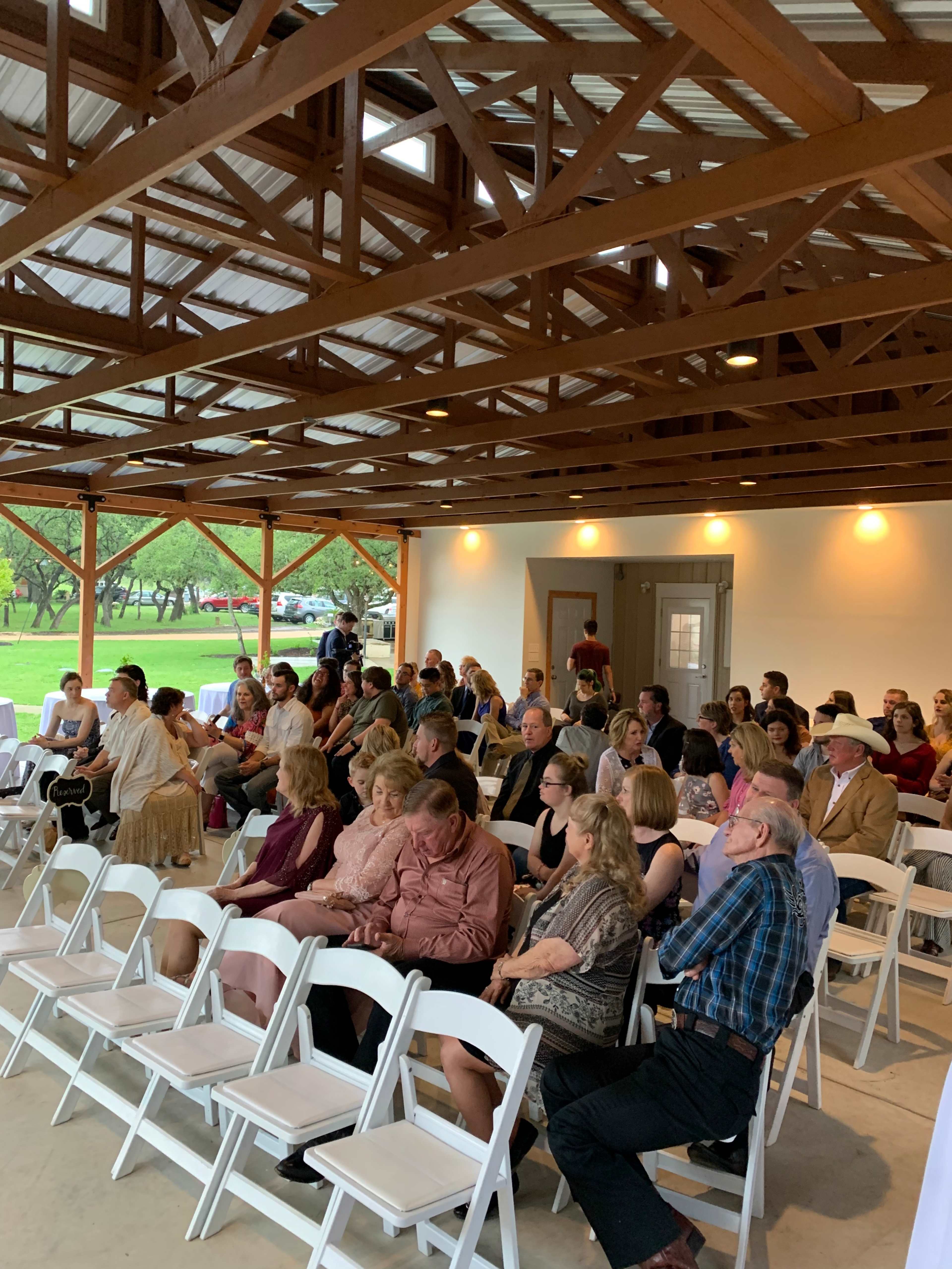 A crowd of people sits in rows of white chairs inside a spacious pavilion with wooden beams, awaiting an event.