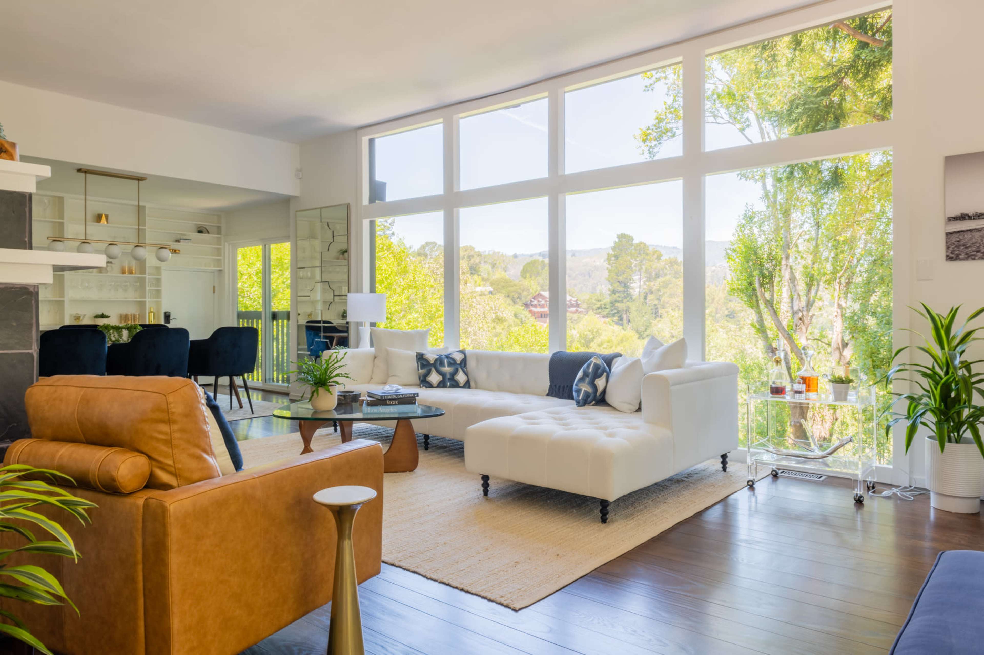 The living room features a large white sectional sofa and floor-to-ceiling windows that provide a view of the trees outside.