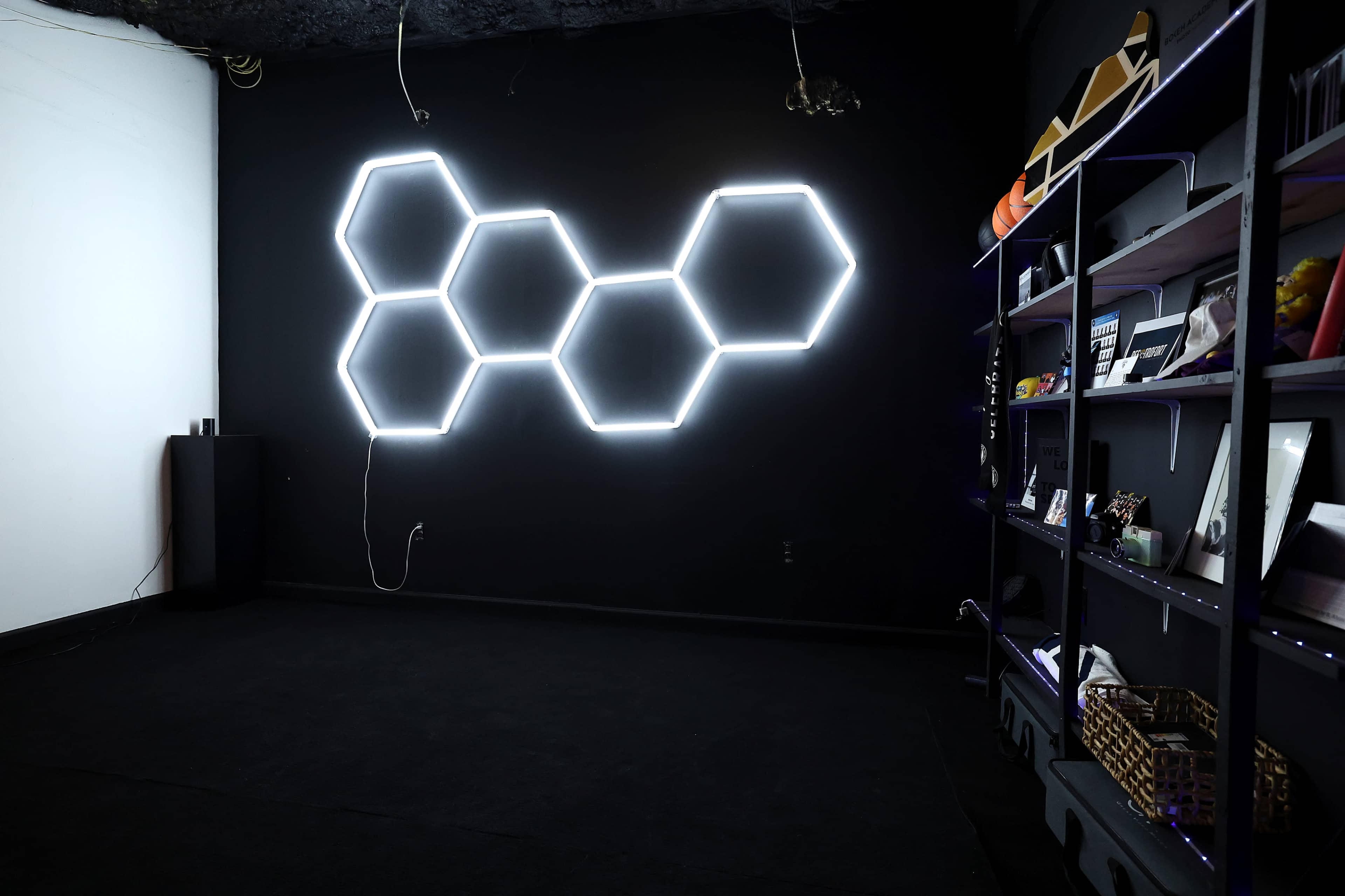 A dark room features a wall with illuminated hexagonal shapes and a shelving unit displaying various items.