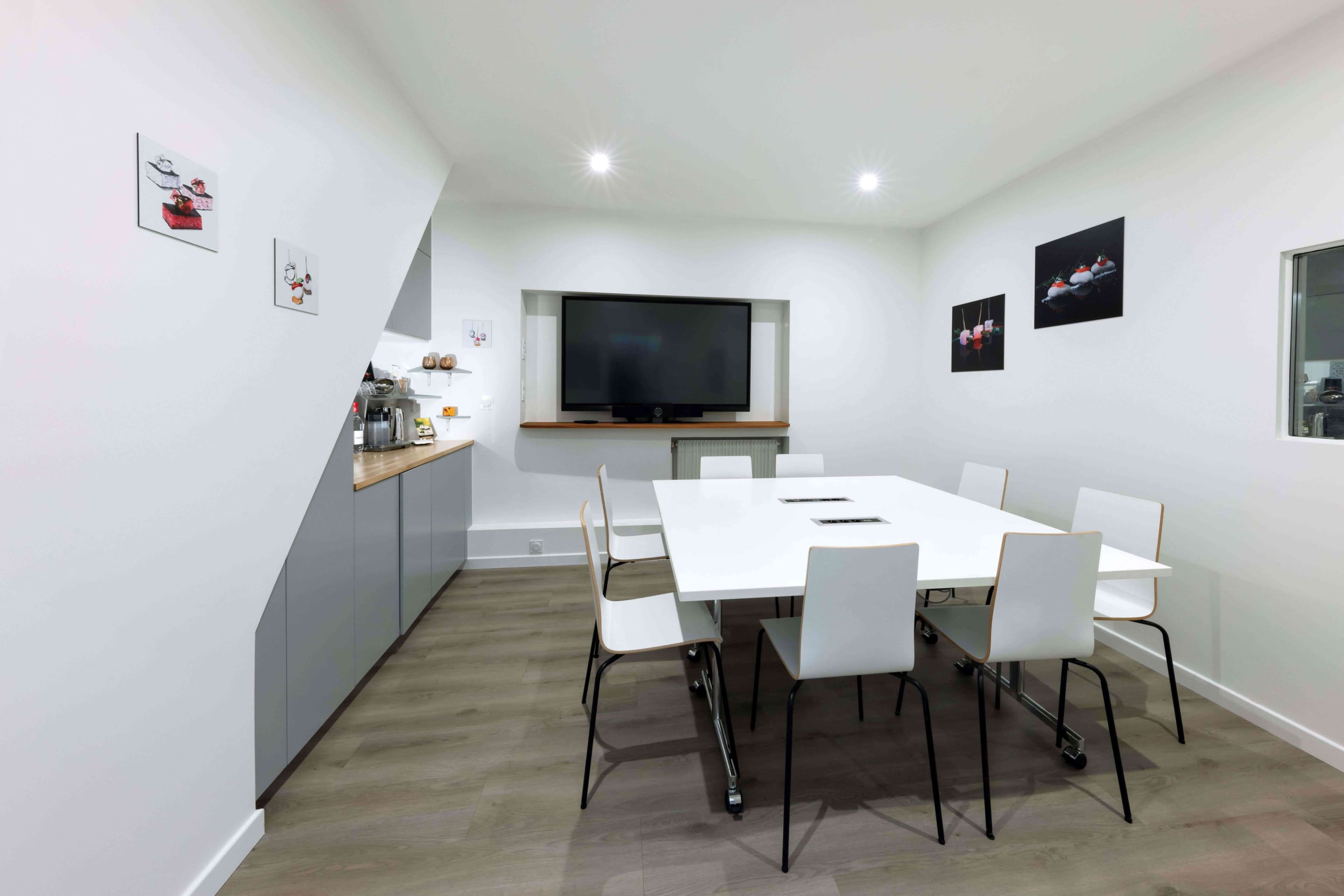 A modern conference room features a large white table surrounded by eight chairs, a wall-mounted television, and framed artwork on the walls.
