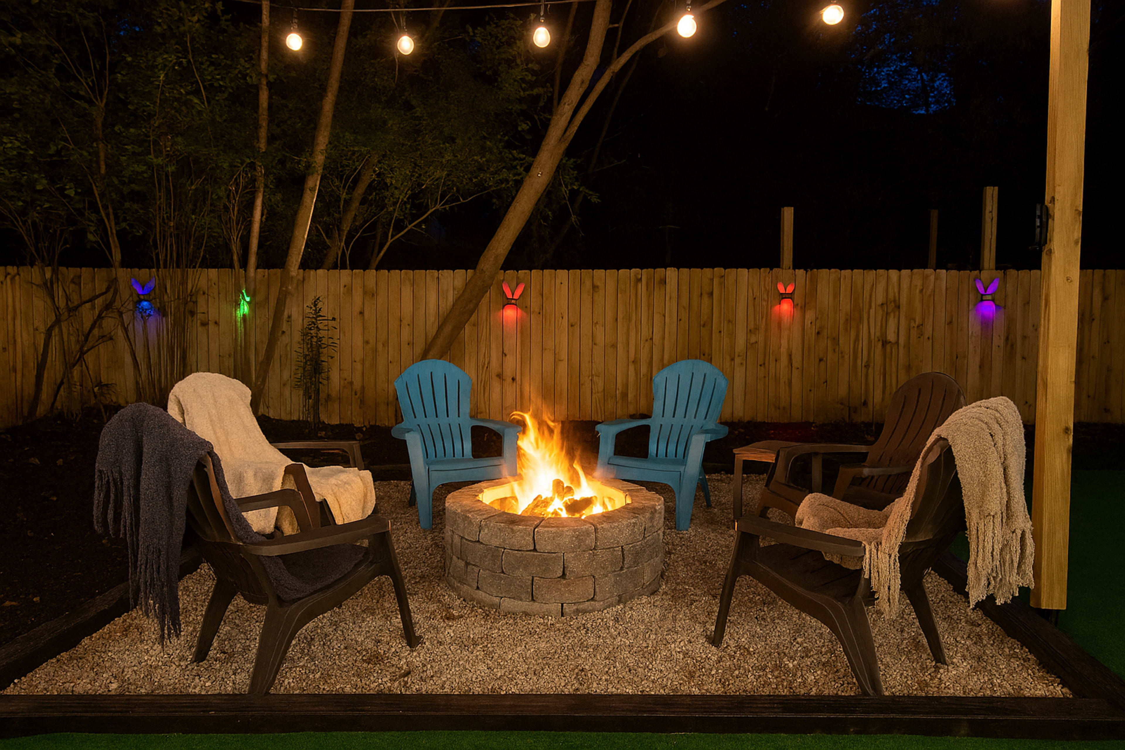 The image depicts a cozy outdoor fire pit surrounded by five colorful adirondack chairs, with string lights illuminating the scene at night.