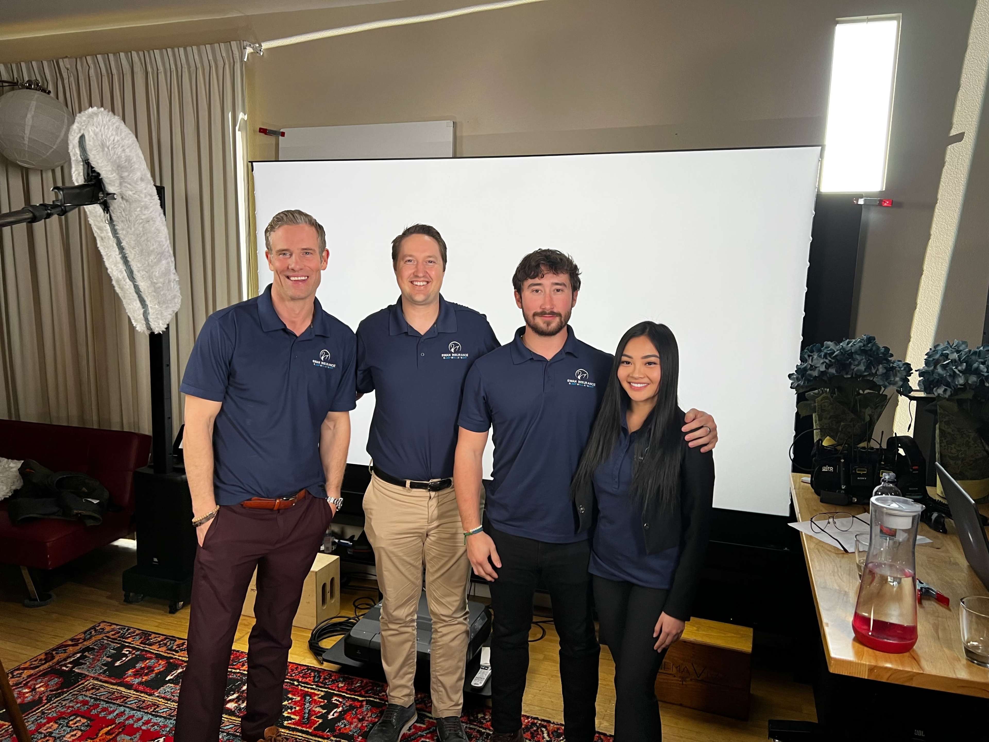 Four people in matching navy polo shirts stand together in a well-lit room with a large screen behind them.