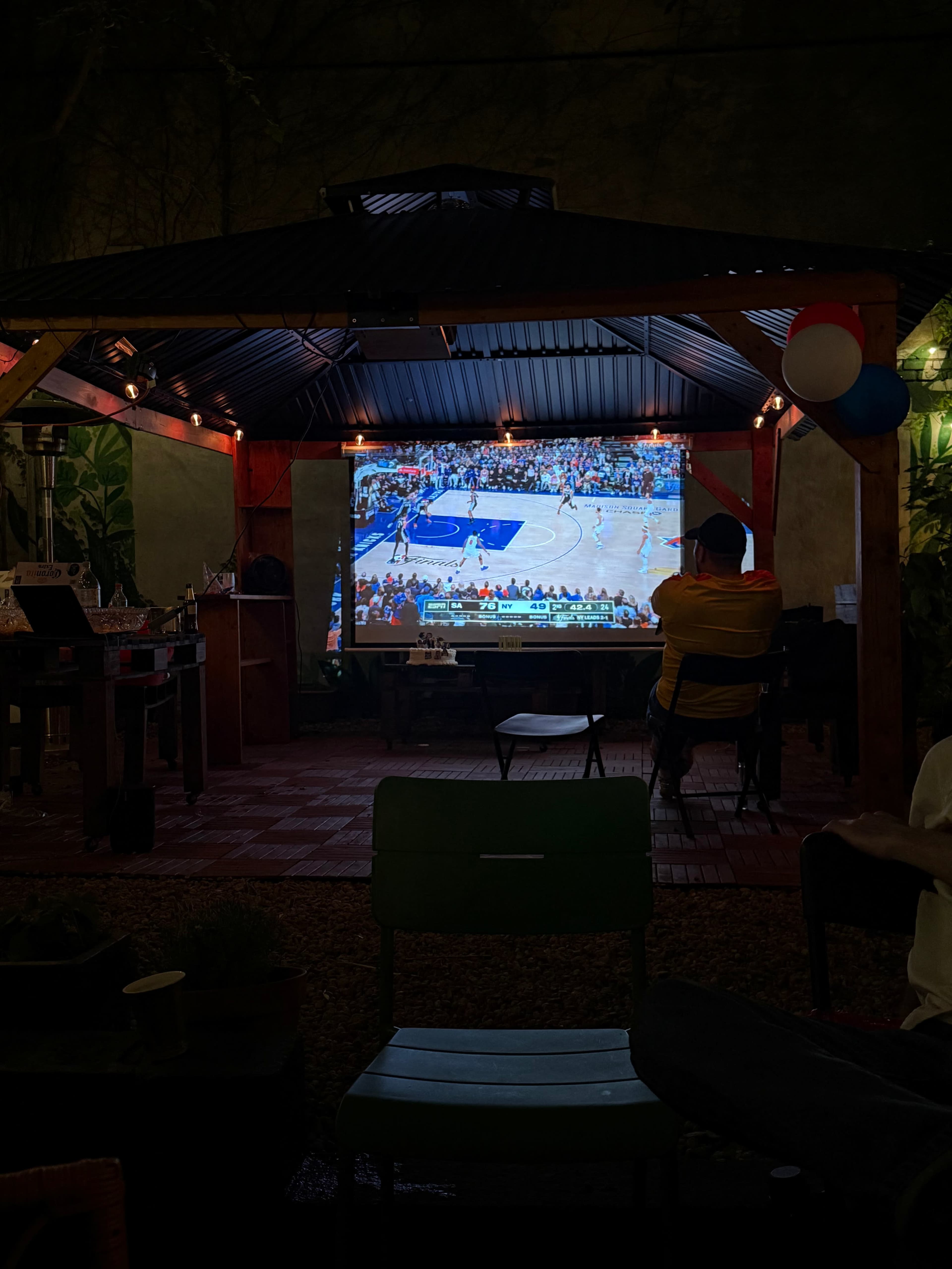 A person sits on a chair in an outdoor shelter watching a basketball game projected on a large screen.