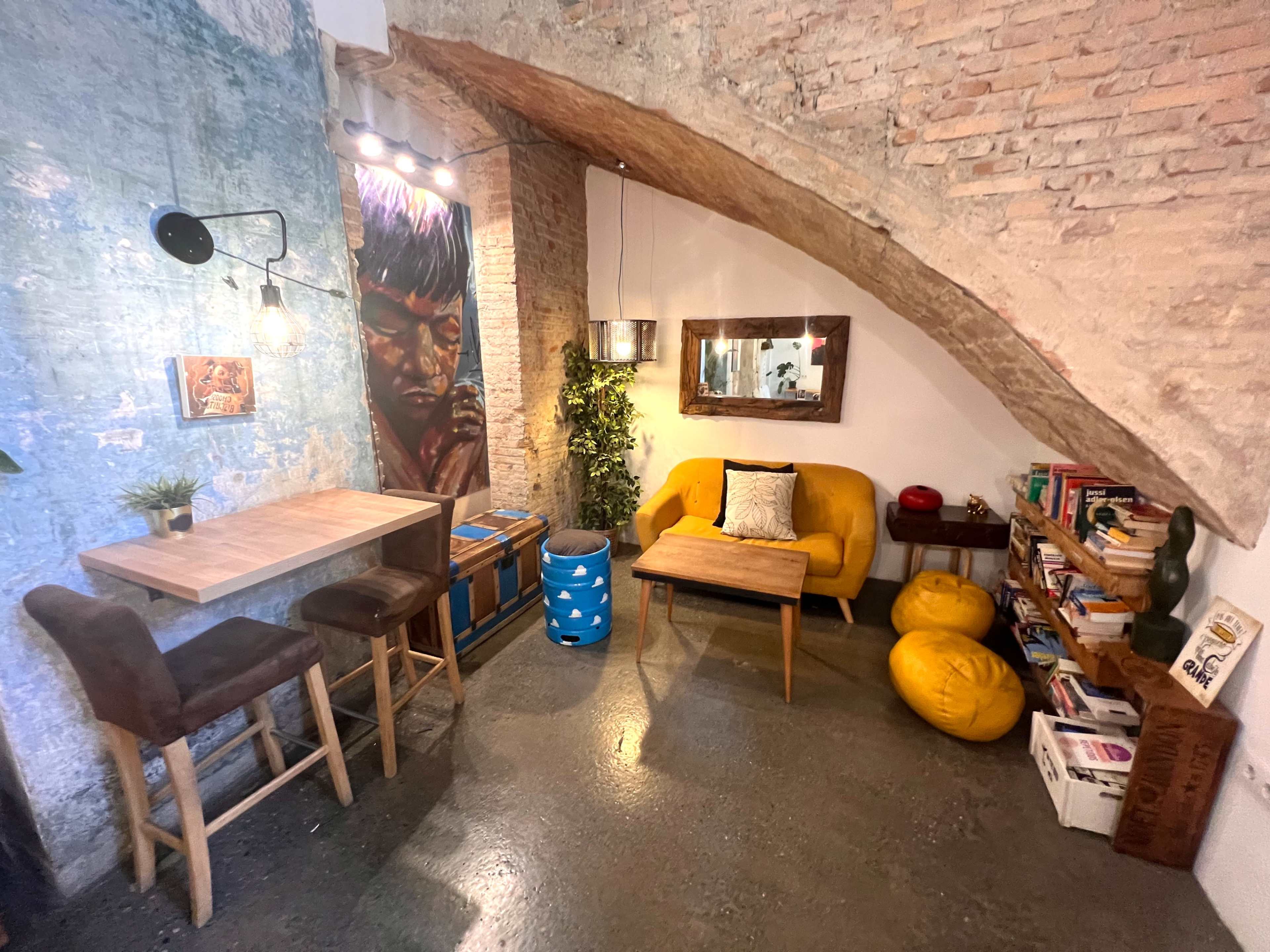 The image shows a cozy corner of a room featuring a yellow couch, a wooden table, and a bookshelf against a textured wall with exposed bricks.
