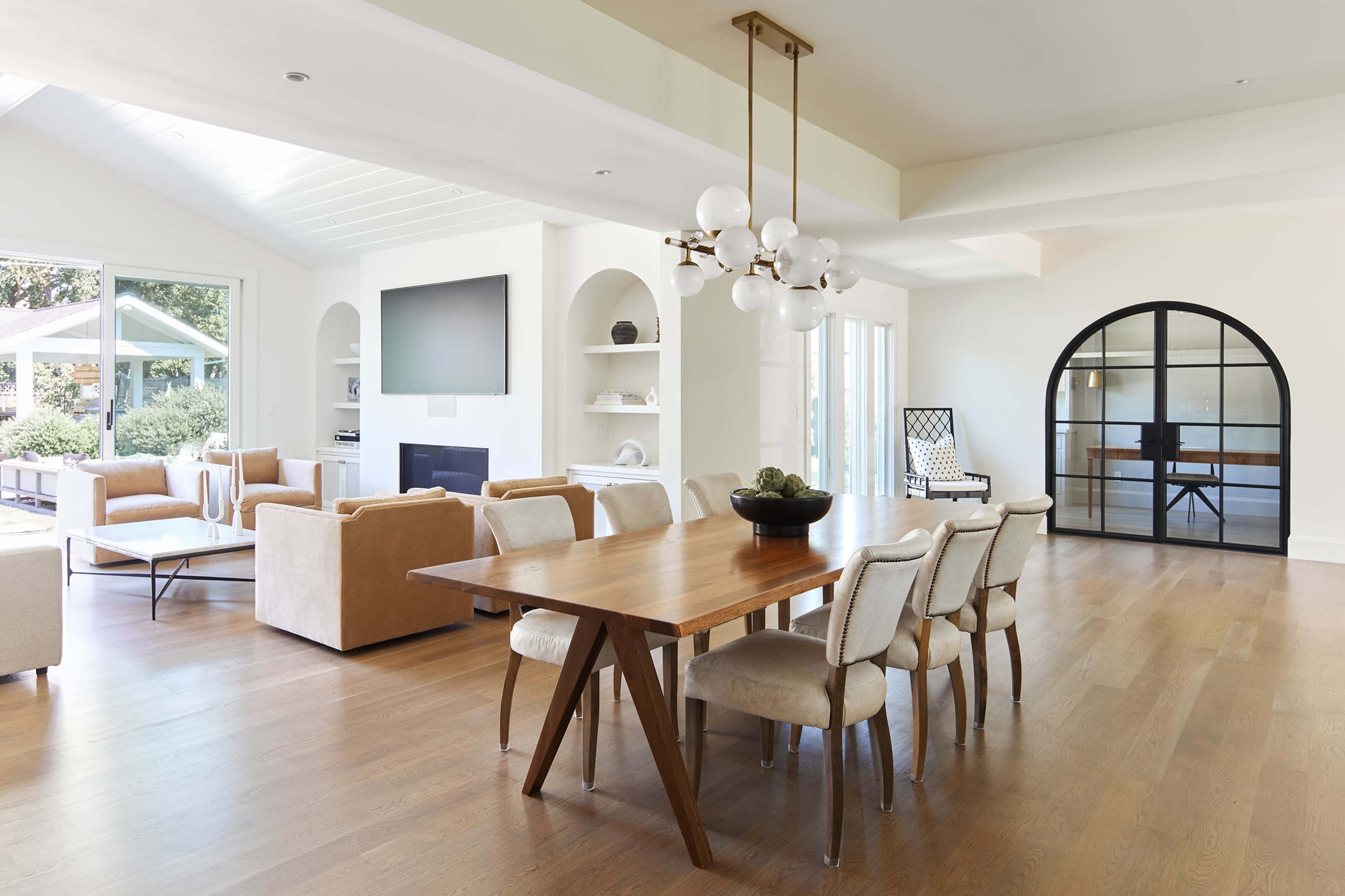 A modern dining area features a wooden table surrounded by upholstered chairs, adjacent to a spacious living area with large windows and a glass door.
