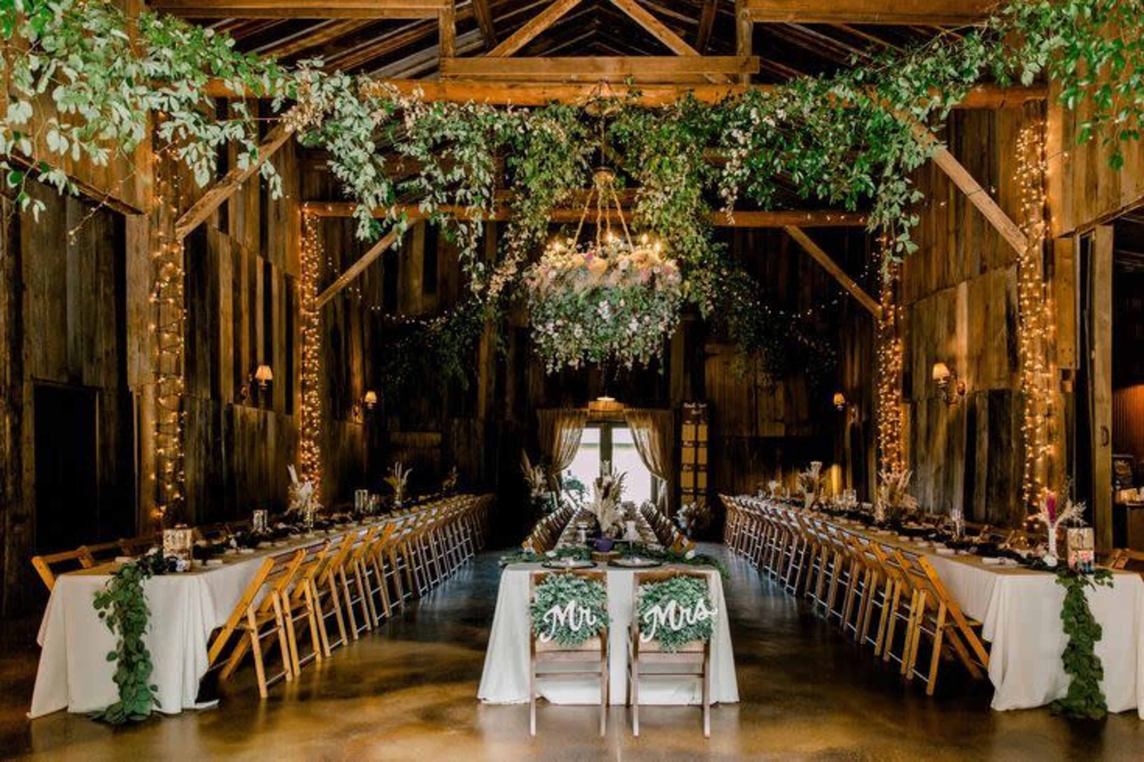 The image shows a rustic barn interior decorated for a wedding reception, featuring long dining tables with floral centerpieces, string lights, and an elegant chandelier surrounded by greenery.