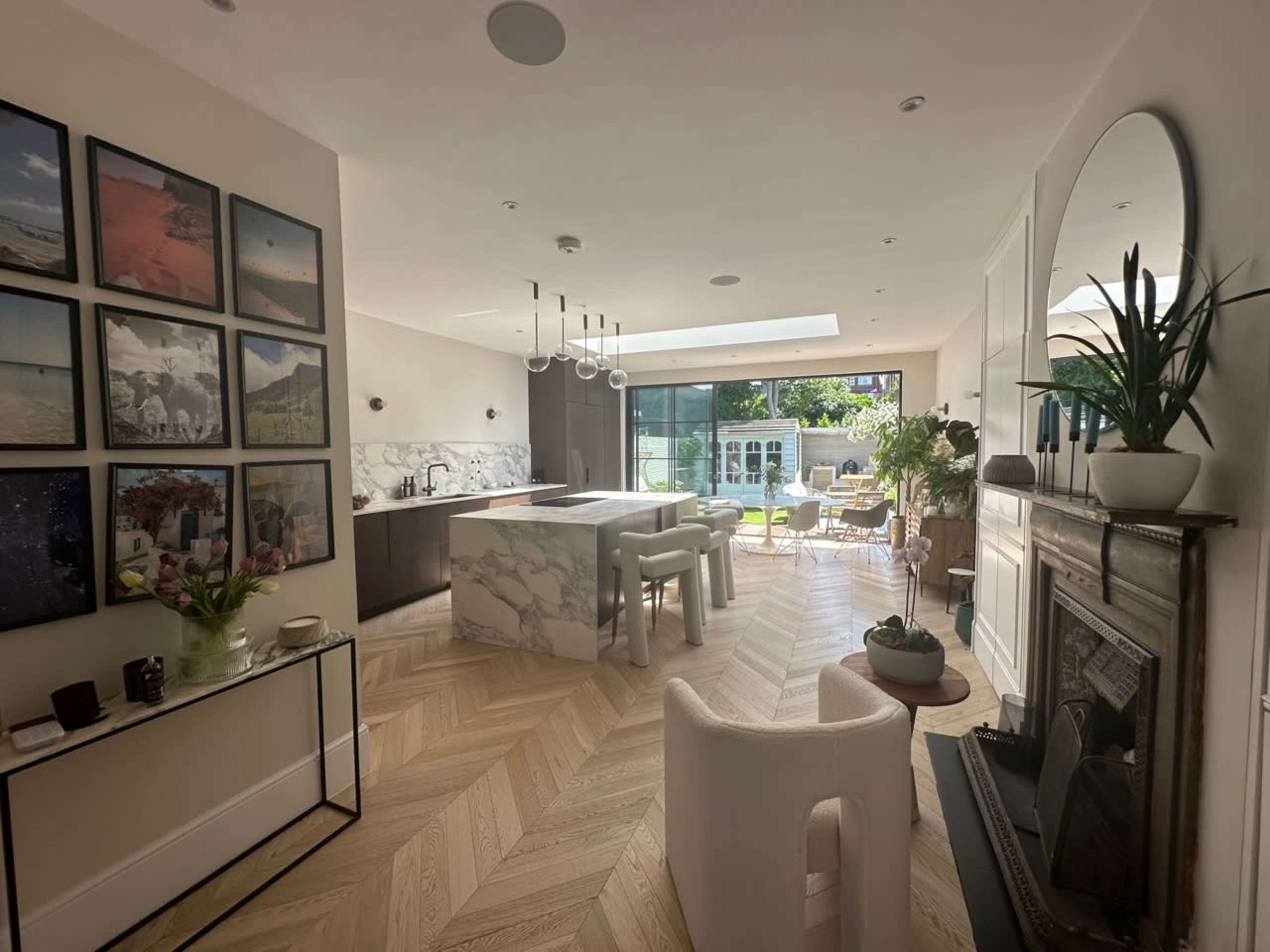 The image shows a modern, open-concept kitchen and living area with marble countertops, a large window leading to an outdoor space, and several decorative photographs on the walls.