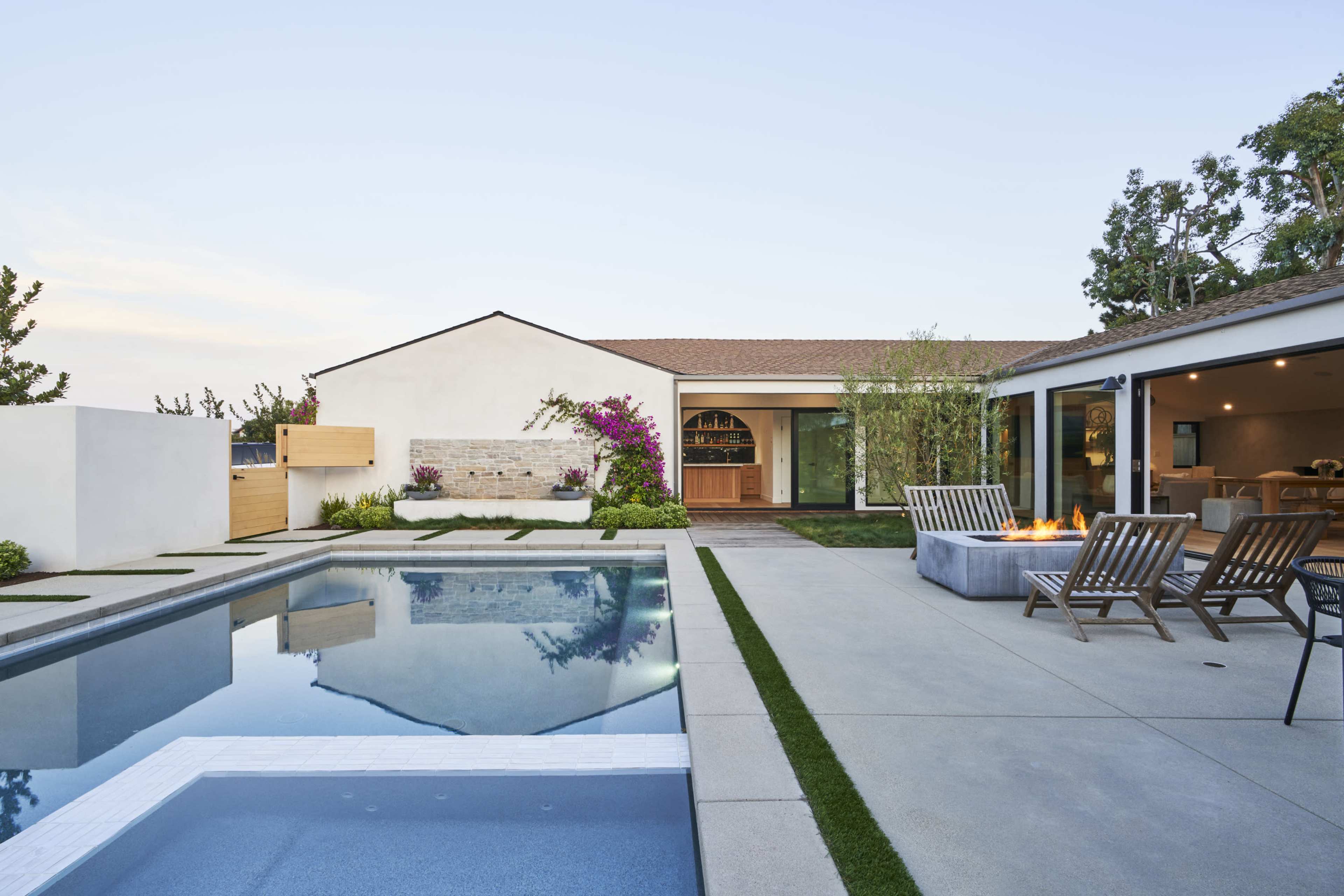 A modern backyard with a swimming pool, patio area, fire pit, and a house featuring large windows and landscaping.