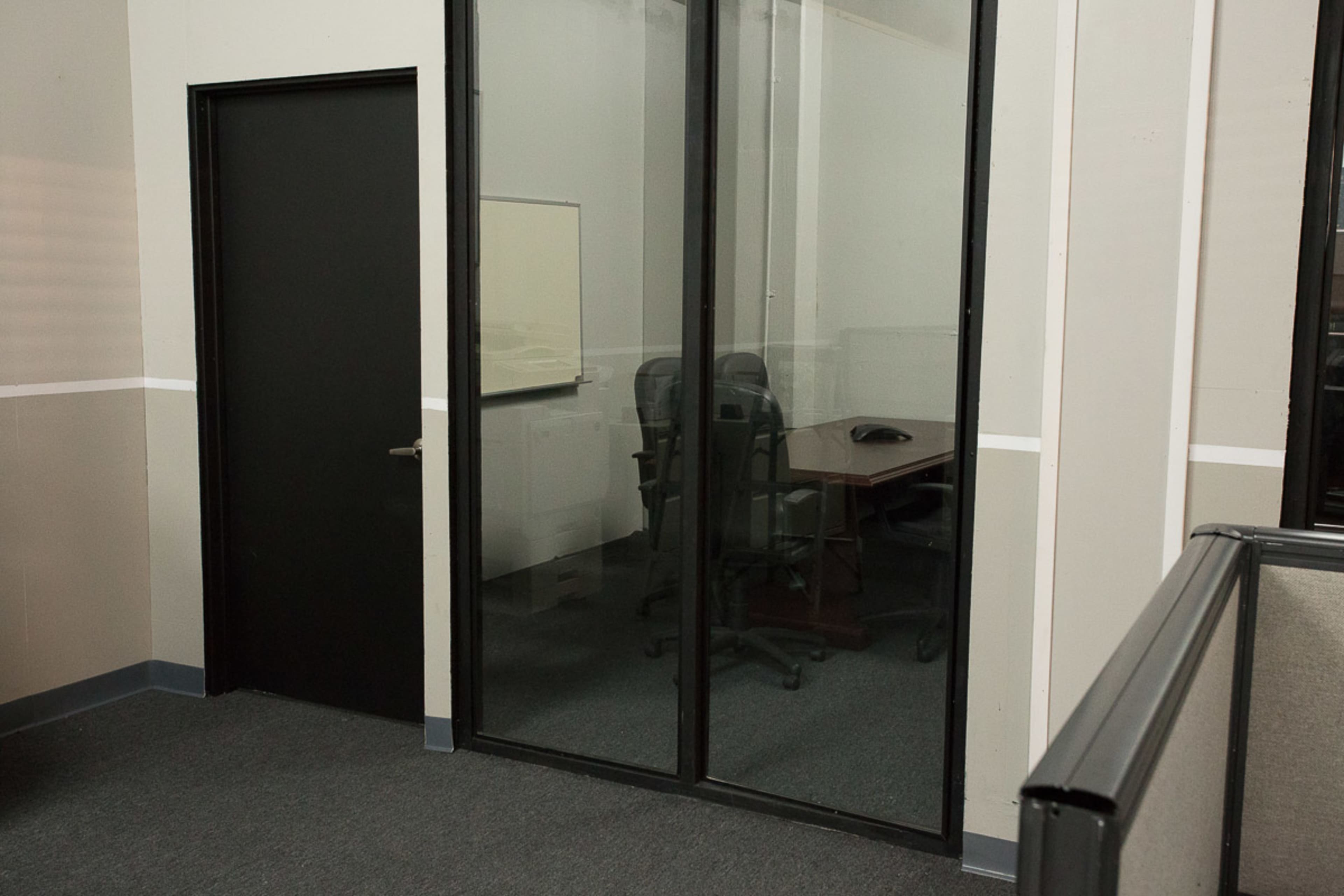 A small conference room with a glass wall contains a table and chairs, adjacent to a black door in an office space.