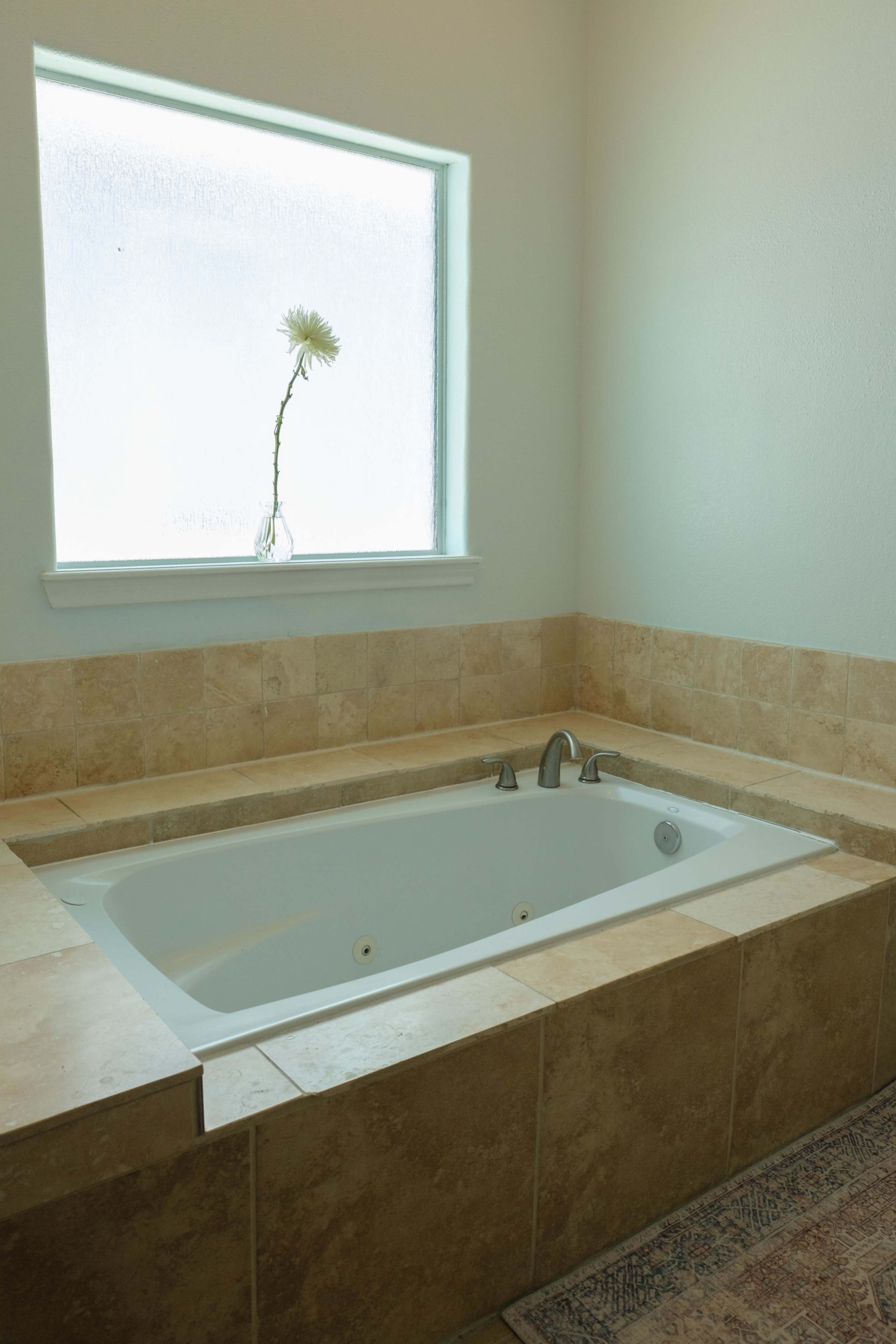 A sunlit bathroom features a white bathtub surrounded by beige tiled walls and a window with a single flower in a vase.
