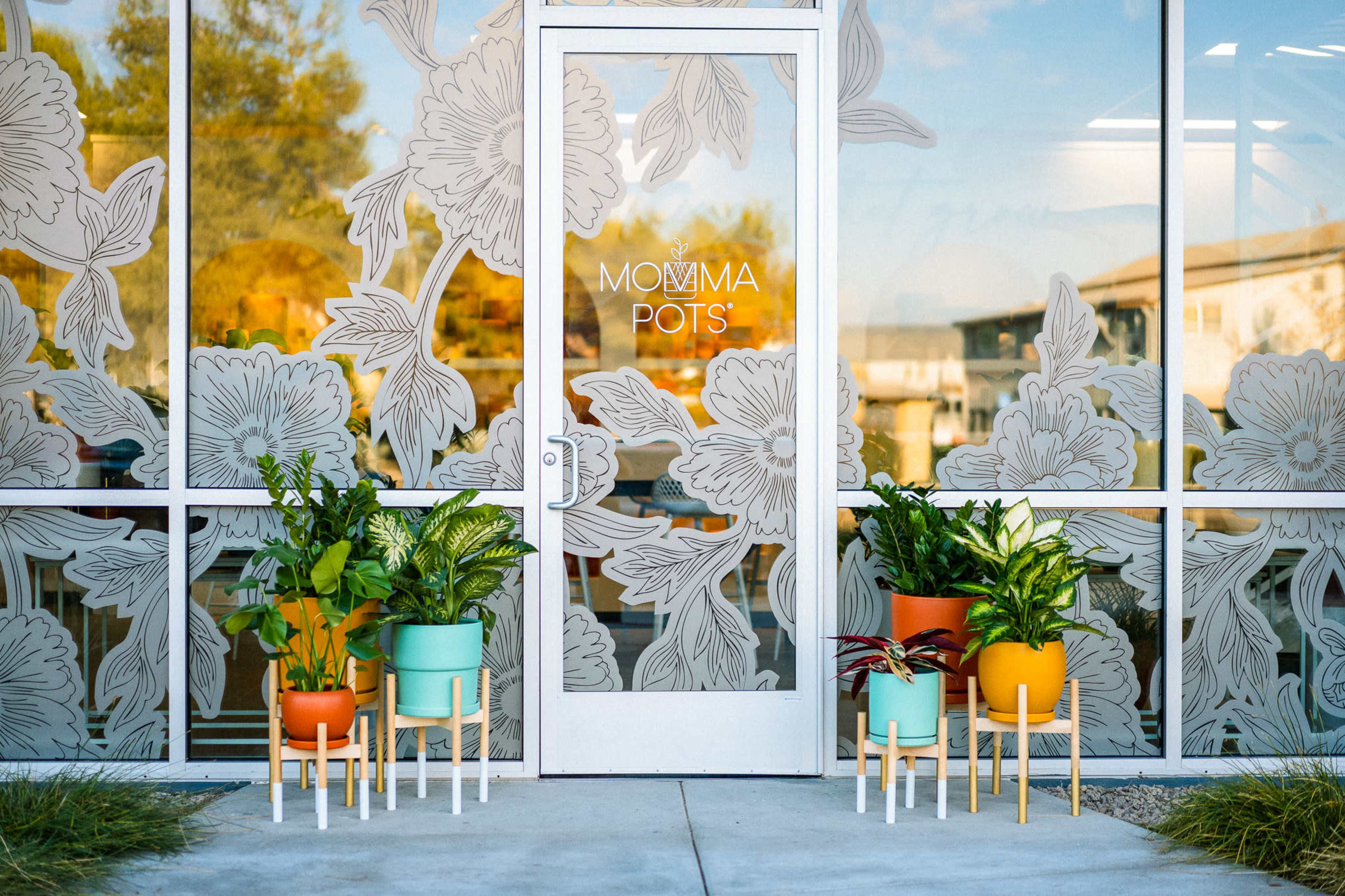 A glass entrance with floral-patterned decals and potted plants in colorful pots arranged outside the door.