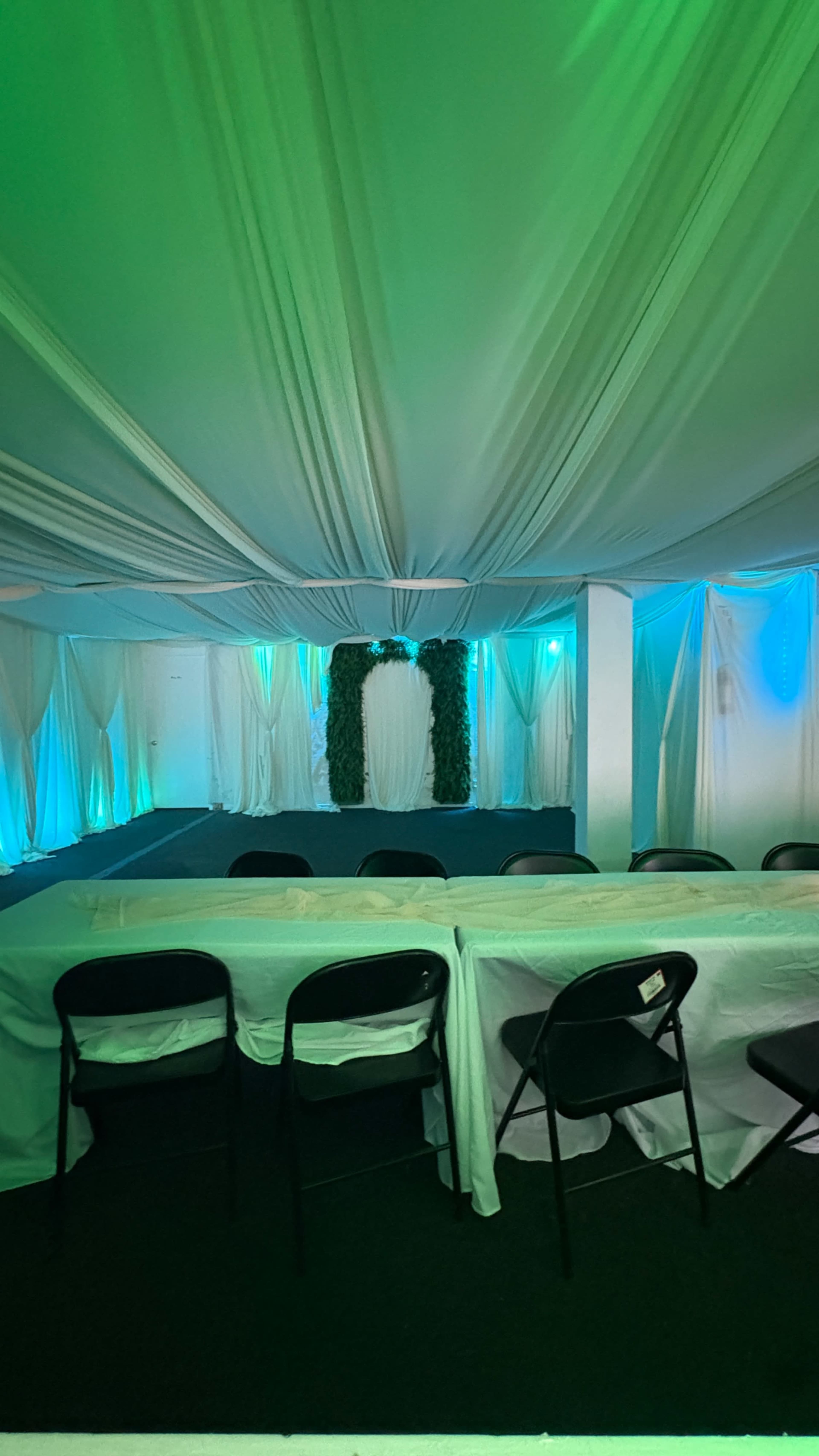 The image shows a decorated event space with draped fabric, green and blue lighting, and a central backdrop featuring a floral arrangement.