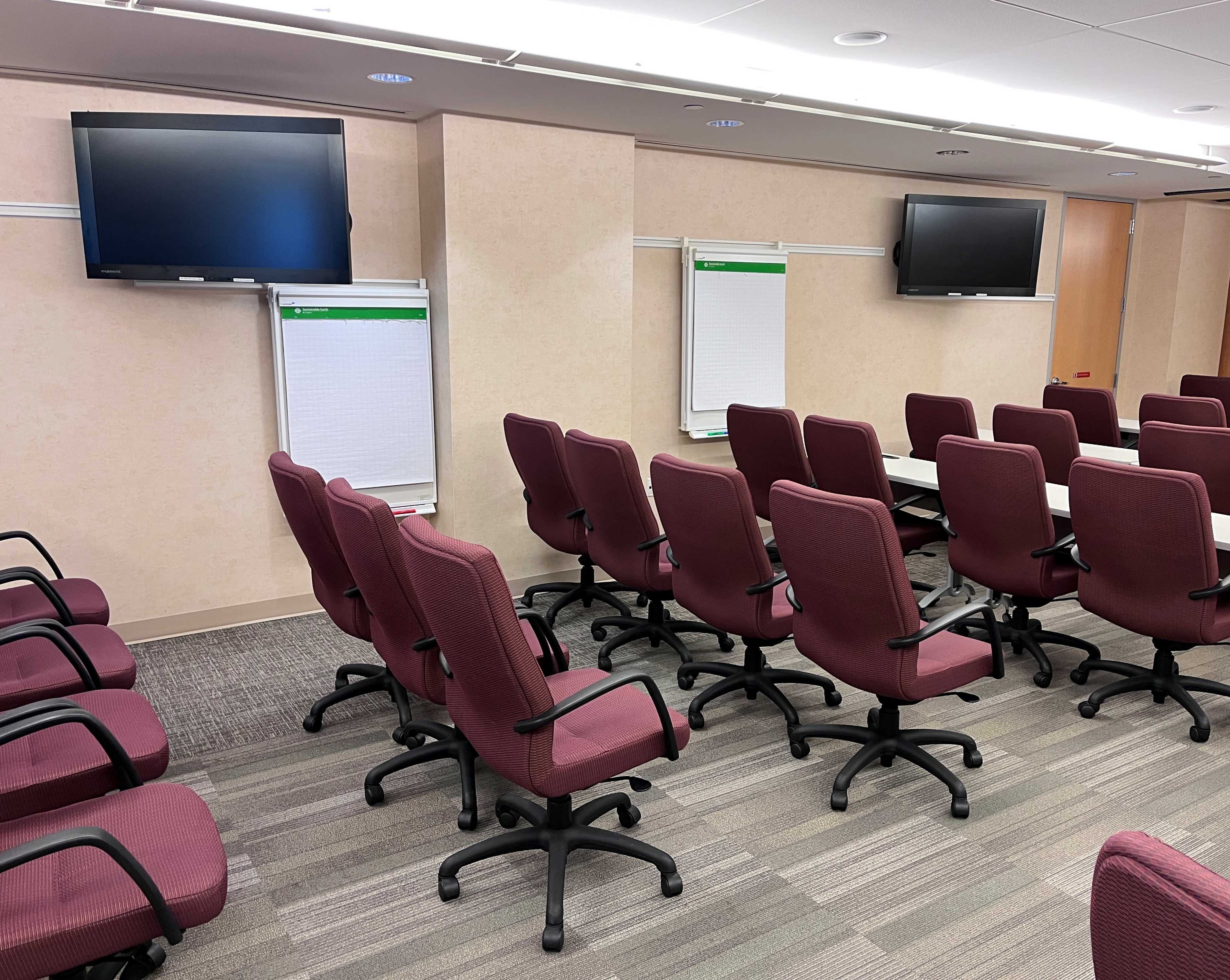 The image shows a conference room with several burgundy chairs arranged around a long table, two monitors mounted on the walls, and whiteboards positioned nearby.