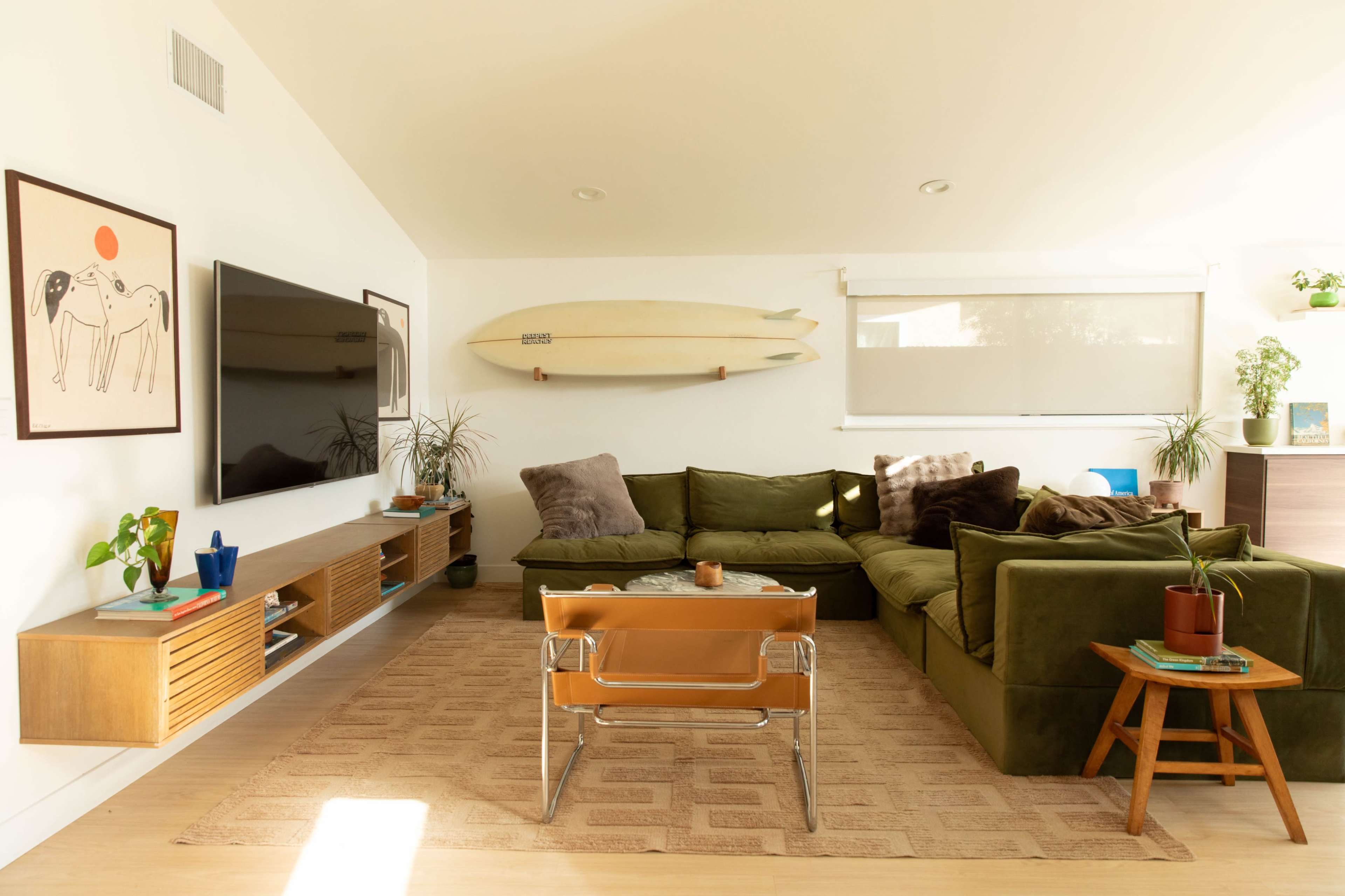 The image depicts a modern living room featuring a green sectional sofa, a wall-mounted TV, surfboards, and various houseplants.