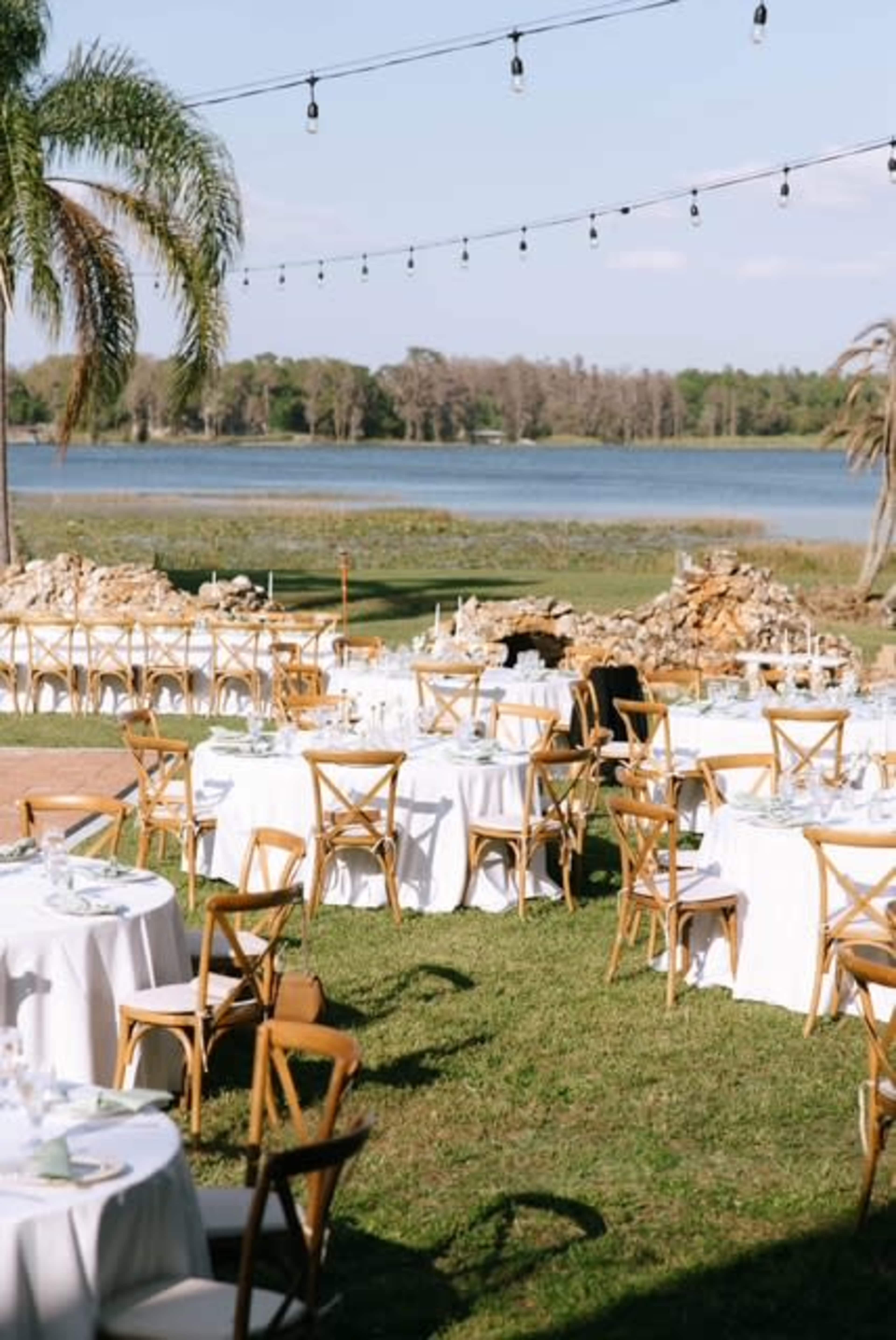 An outdoor event venue features round tables with white tablecloths set up on a lawn next to a tranquil body of water, framed by greenery and string lights overhead.