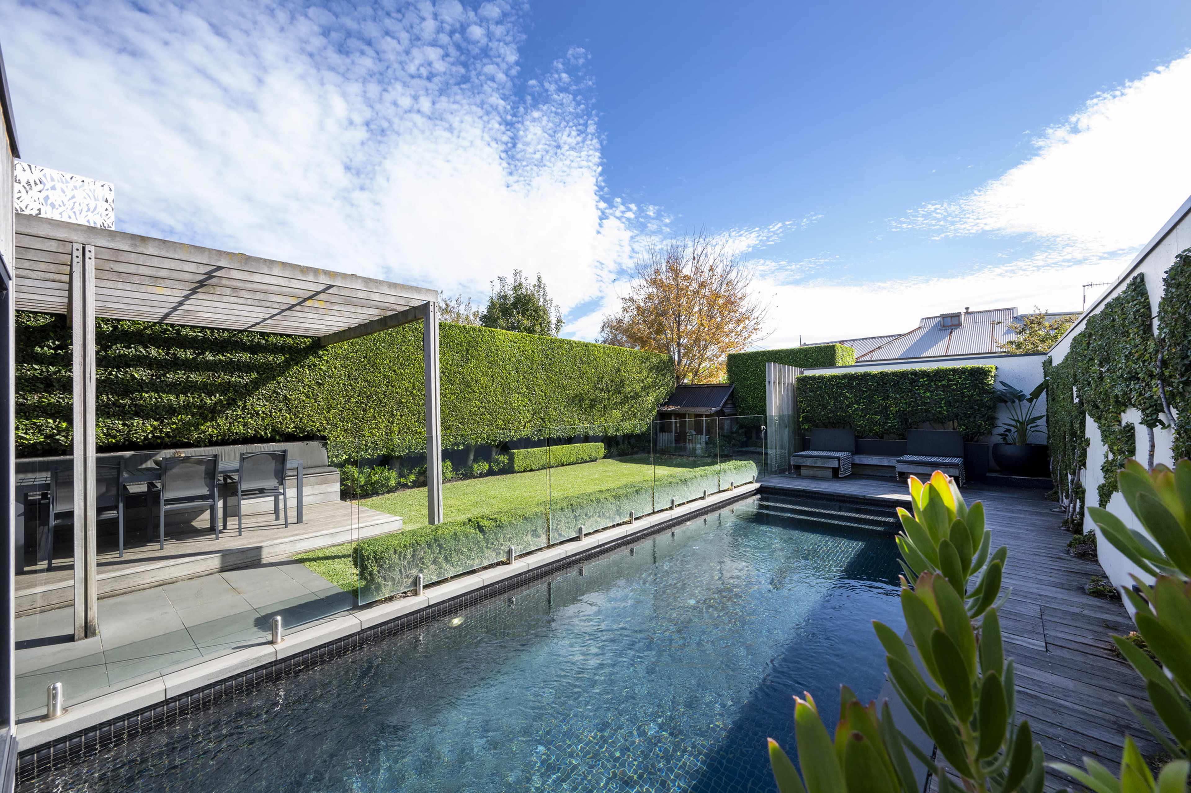 The image shows a contemporary backyard with a swimming pool surrounded by landscaped greenery and a covered outdoor dining area under a wooden pergola.