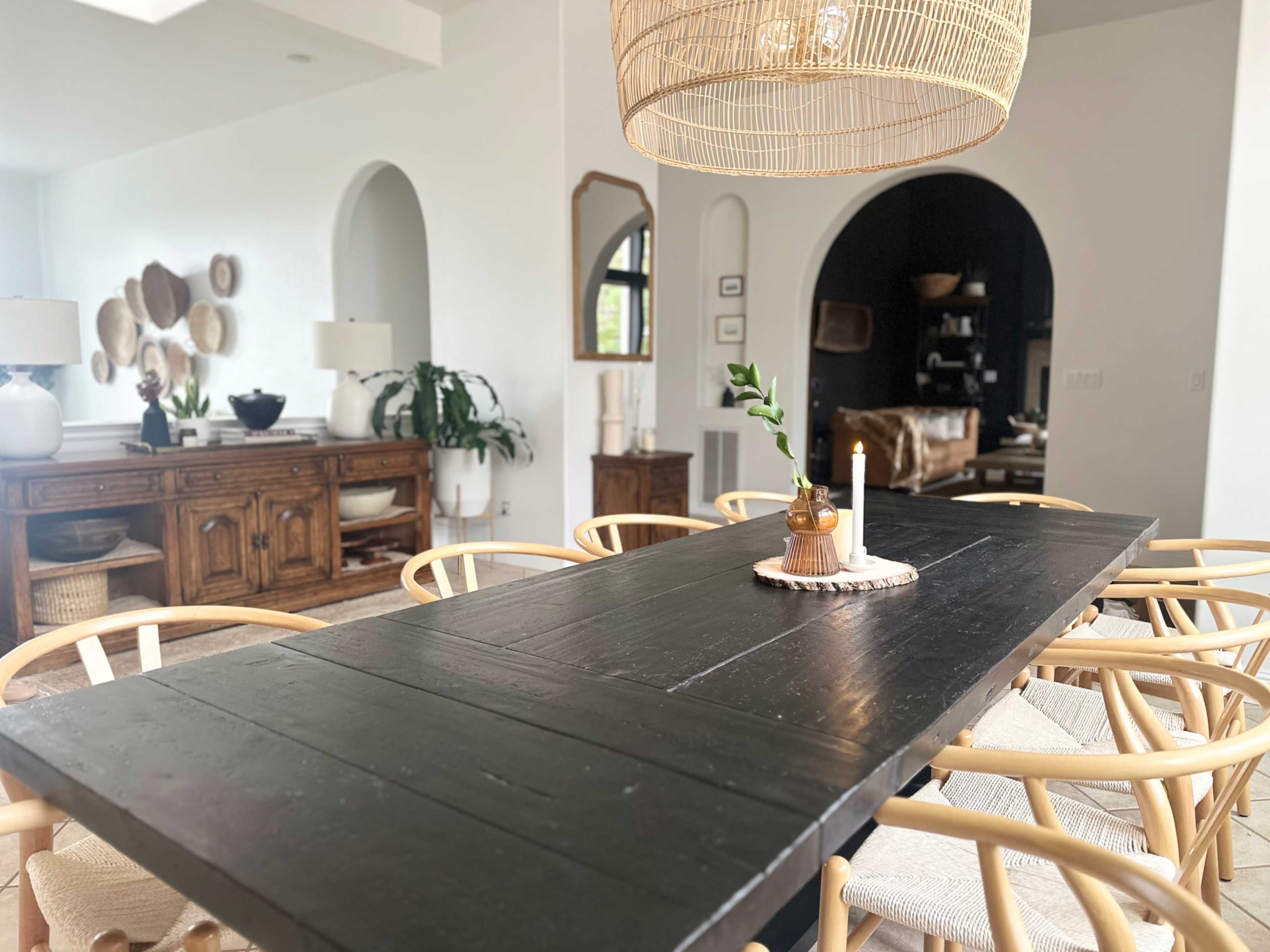 A large black dining table is set in a well-lit room, surrounded by light wooden chairs, with a decorative centerpiece and a woven lantern hanging above.