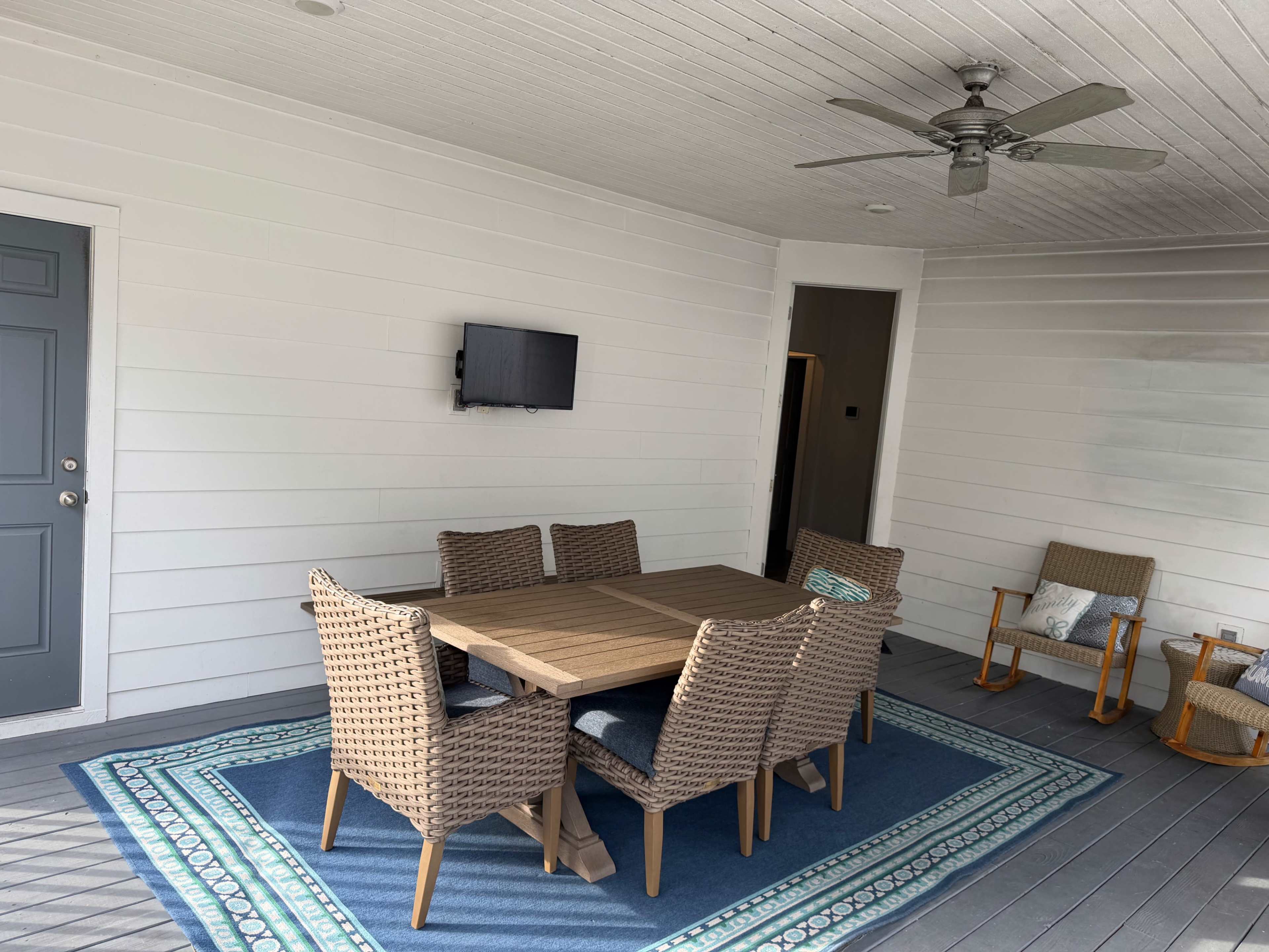 A patio area features a wooden dining table surrounded by six wicker chairs, with a television mounted on the wall and a rocking chair positioned nearby.