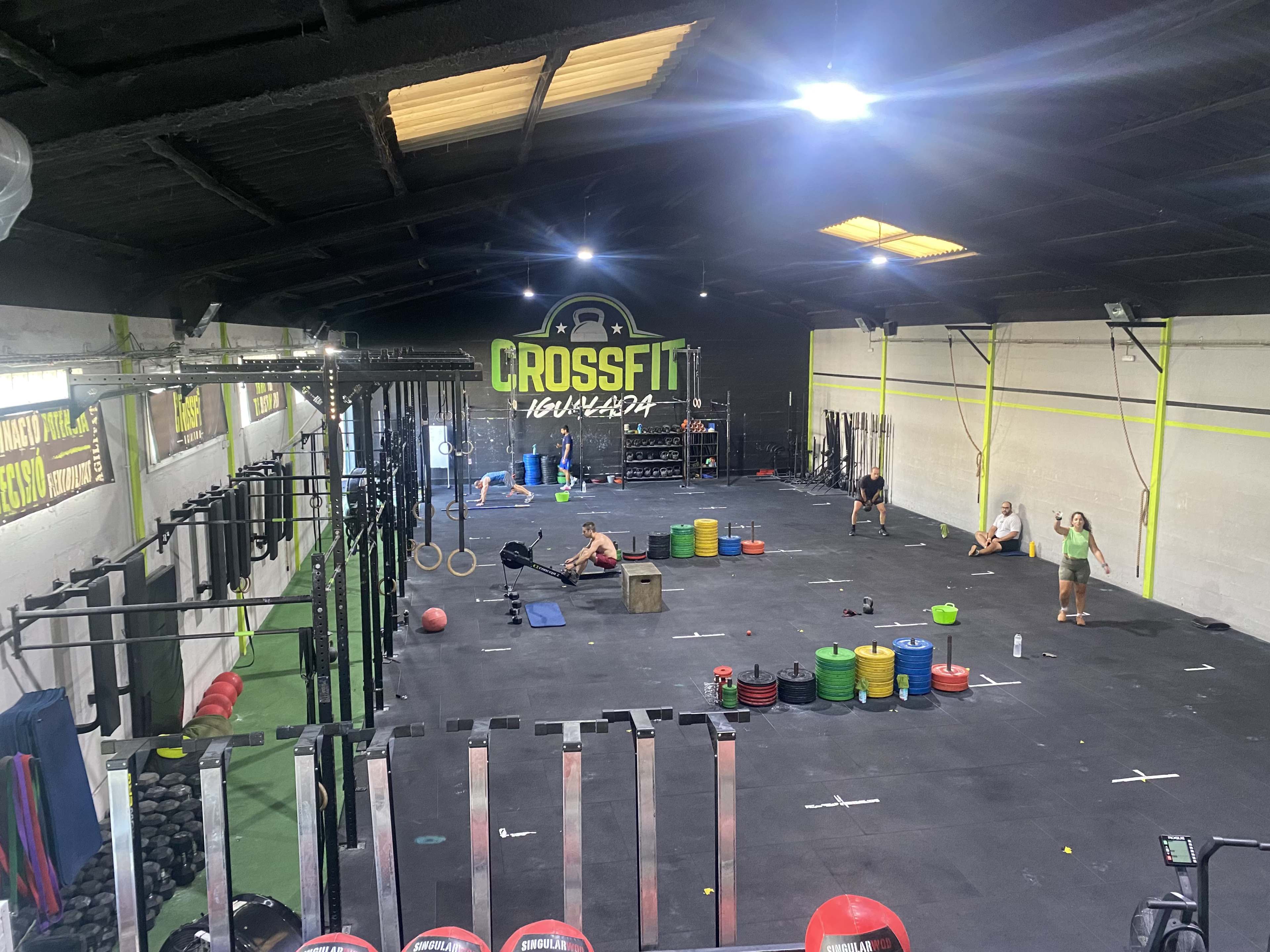 A spacious CrossFit gym features various weightlifting equipment, exercise mats, and several individuals engaged in workouts.