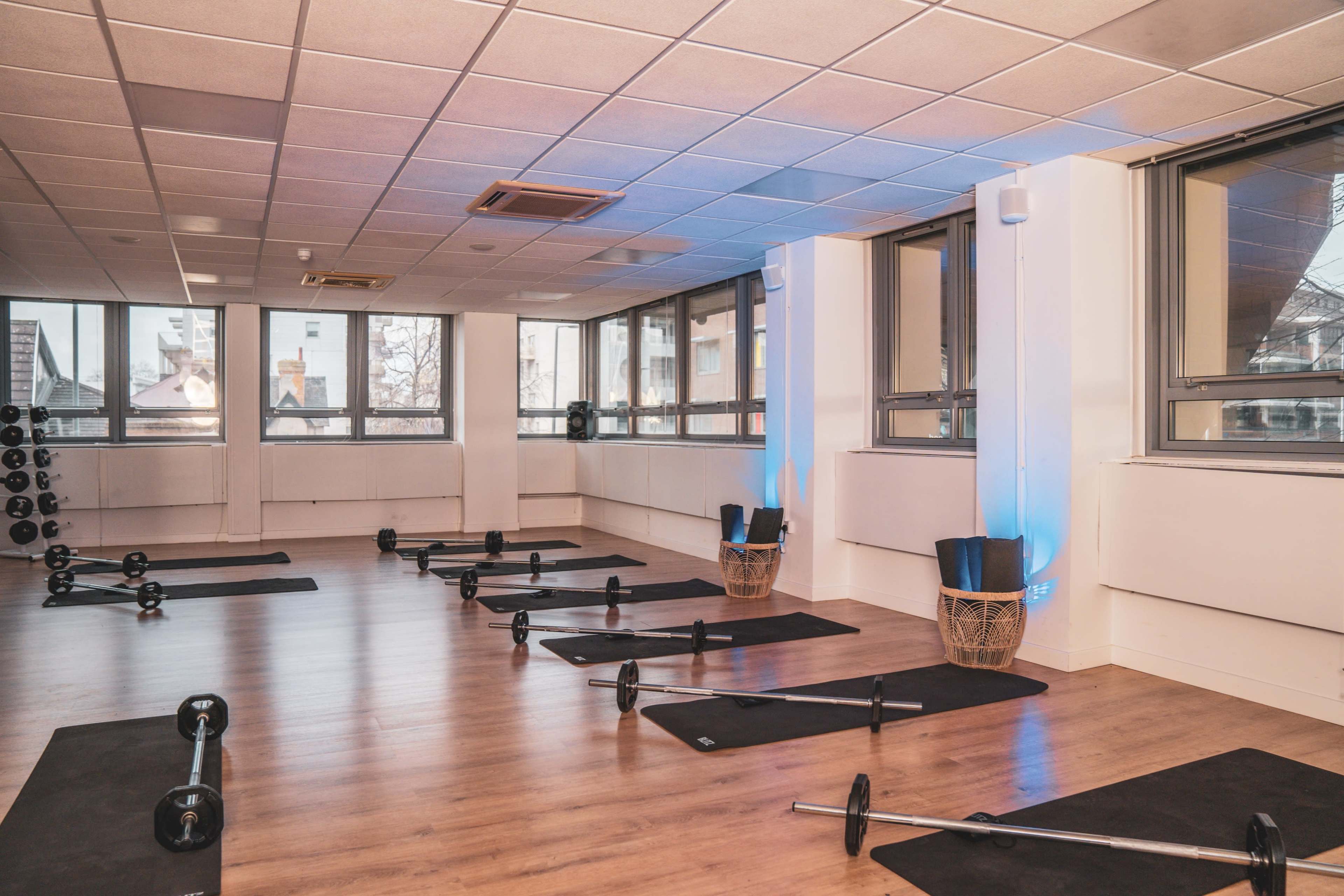 The image shows an empty fitness studio with wooden flooring, exercise mats, and barbell weights arranged neatly.