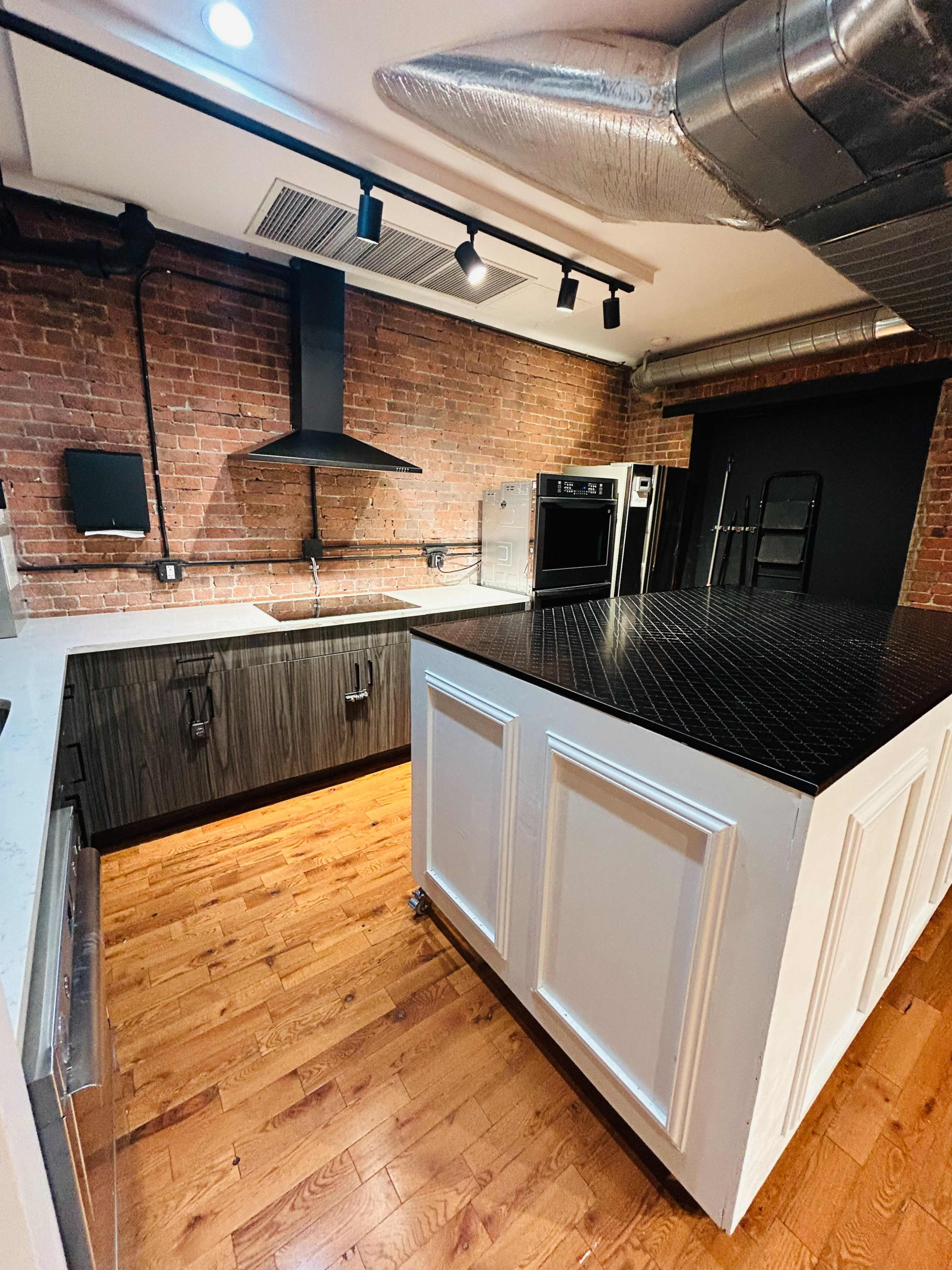 The image shows a modern kitchen with a large central island featuring a dark countertop, surrounded by a blend of exposed brick and sleek cabinetry.