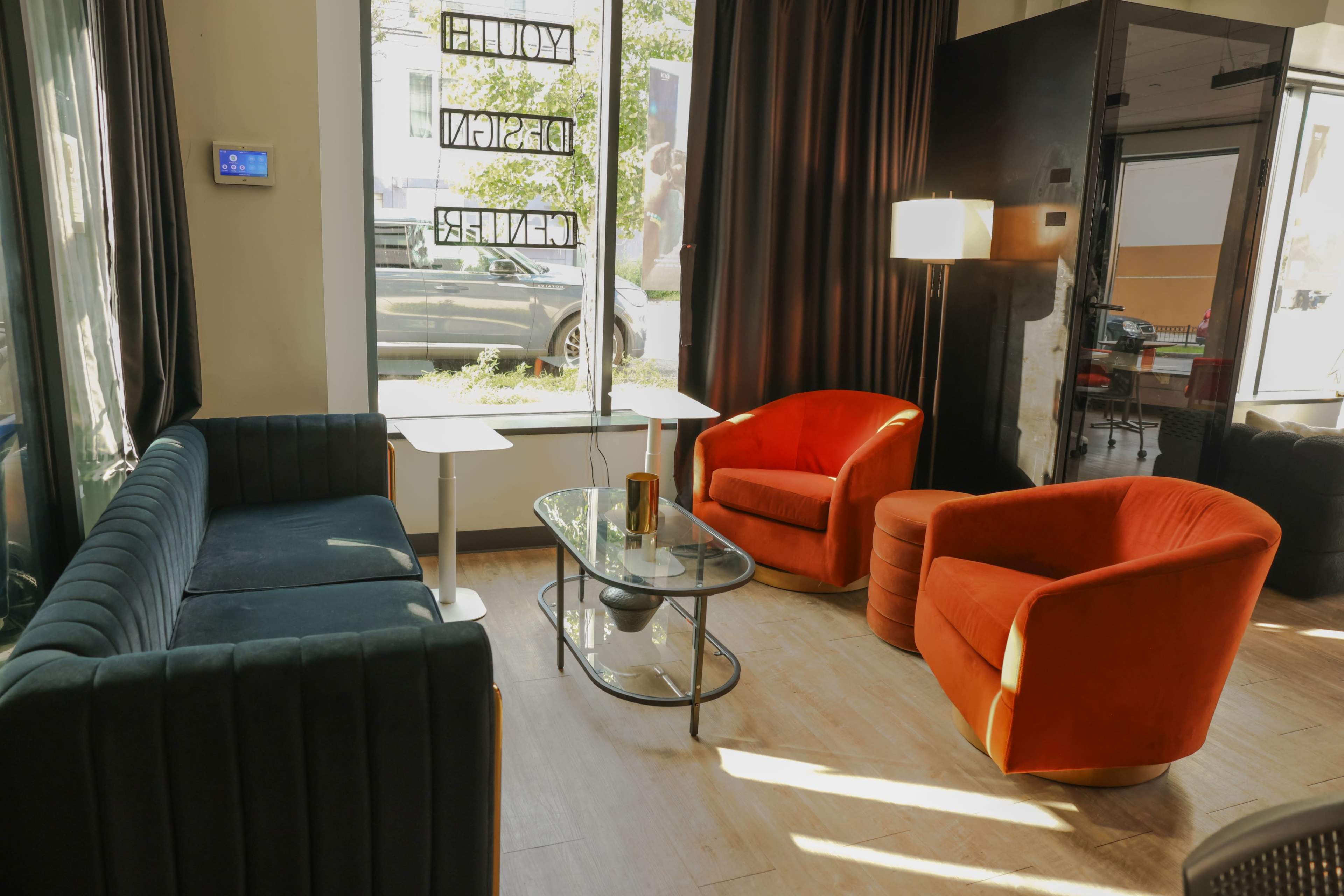The image shows a cozy interior space with two orange armchairs, a green sofa, a glass coffee table, and natural light streaming through large windows.
